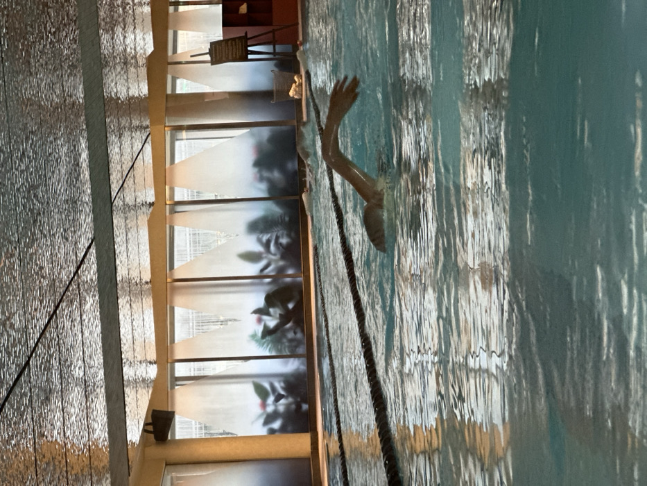 The image displays an indoor swimming pool, identified as the Bank/Vaults pool at The Ned in the City of London, United Kingdom. A lone individual is captured mid-stroke, with their head and an extended right arm visible above the rippling turquoise water, indicating a freestyle swimming motion. Black lane ropes segment the pool horizontally. In the background, a series of large windows feature distinctive frosted panels with dark, silhouetted tropical plant designs, allowing diffused light from the exterior urban environment to enter. The ceiling above is textured and highly reflective, showing distorted reflections of the surroundings. To the right of the pool, an easel supports a dark display board, and a wicker basket containing rolled white towels is placed beside it. A dark, rectangular speaker is mounted on the left wall above the windows. The scene is evenly lit.