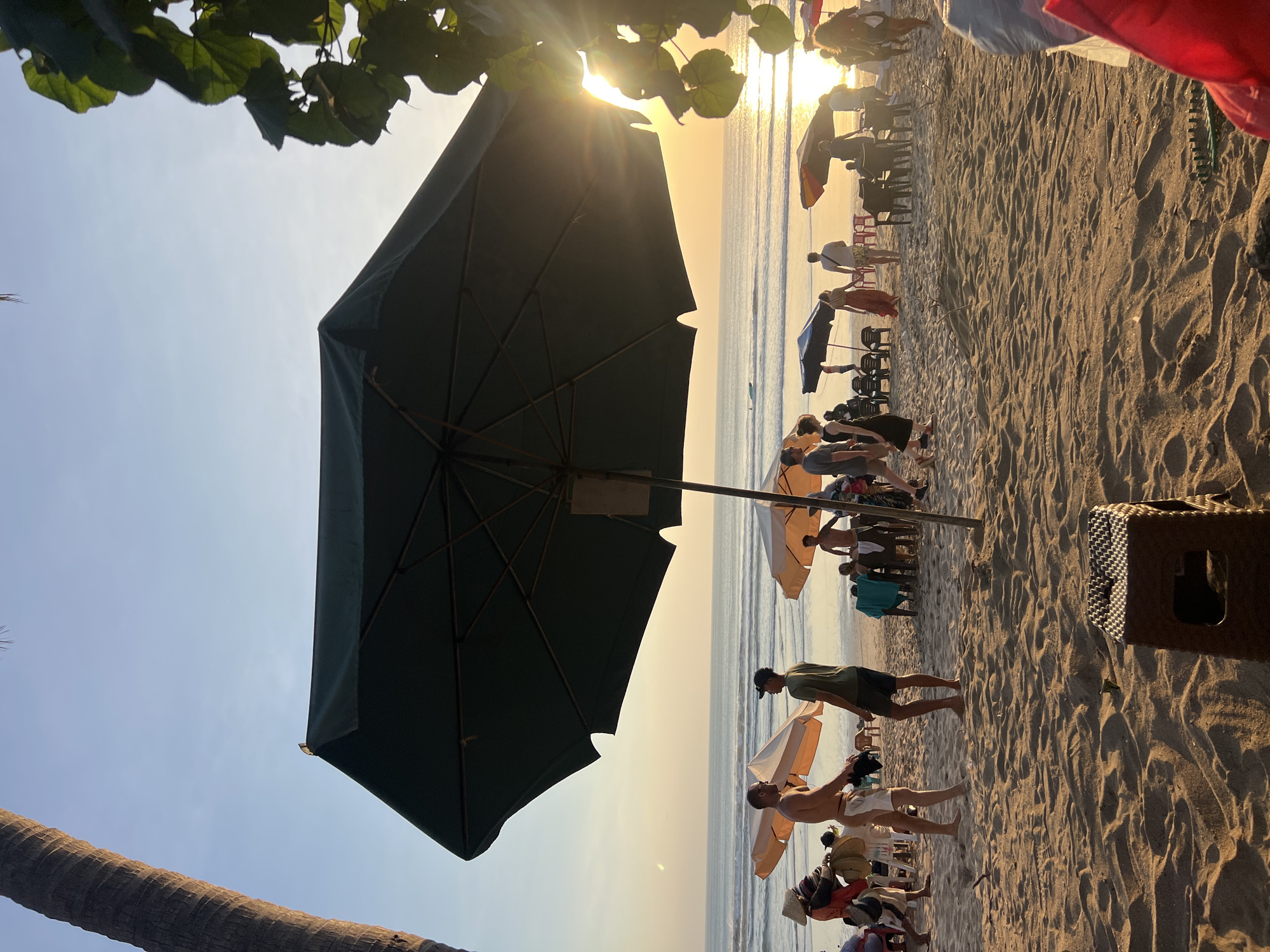 This image depicts a beach scene in Kuta, Indonesia, during sunset. The foreground is dominated by textured sand, with a small brown plastic stool in the lower right corner. A large dark green umbrella casts a shadow over a section of the beach, with its spokes visible. Several people are scattered across the sand, some walking, others standing or sitting under umbrellas.
In the mid-ground, a group of people are gathered near some orange and black umbrellas, with chairs and tables set up, suggesting a dining or lounging area. Two men are the most prominent figures in this area, one with fair skin and shorts, the other in a green t-shirt and shorts. A woman wearing a straw hat and traditional clothing is visible to the left, interacting with others.
The background shows the ocean with gentle waves and the sun setting, casting a warm golden glow on the water and sky. The horizon is clear, with a faint hint of a boat in the distance. A palm tree trunk is visible on the far left, and lush green foliage from another tree encroaches on the upper right corner, framing the sunset. The overall atmosphere is relaxed and lively, characteristic of a popular tourist beach.