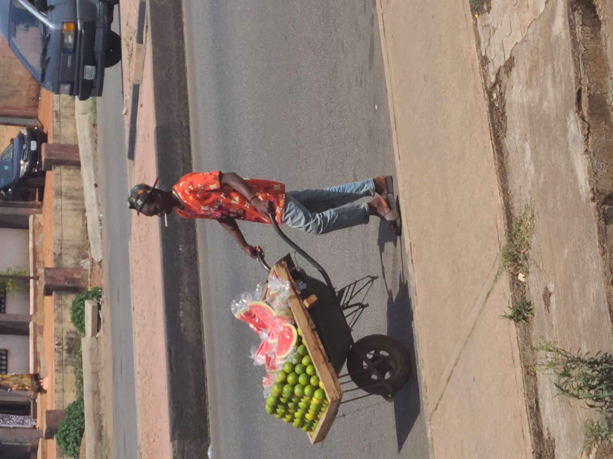 This image depicts a sunny street scene in Jos, Nigeria, featuring a street vendor pushing a fruit cart. The photo is taken from a slightly elevated perspective, looking down onto the road and sidewalk.
The main subject is a person, likely a male, with dark skin, wearing a vibrant orange patterned short-sleeved shirt, light blue jeans, and dark open-toed sandals. A dark hat or head covering is on their head. They are actively pushing a wooden, two-wheeled cart laden with produce. The cart contains large slices of watermelon, some individually wrapped in clear plastic, prominently displayed at the top. Below the watermelon, numerous small green fruits, most likely limes, are neatly stacked in rows.
The setting is an urban street. To the left of the vendor, a paved asphalt road extends into the background, where two vehicles are visible: a dark blue Toyota van with "TOYOTA" visible on its front, and another dark car behind it. On the right side of the vendor, a concrete sidewalk runs parallel to the road. The sidewalk shows signs of wear, with cracks, dirt, and patches of sparse vegetation along its edge. Beyond the road on the left, multi-story buildings with concrete facades and some greenery are visible.
The clear, bright sunlight indicates it is daytime, casting distinct shadows from the vendor, the cart, and its wheel onto the road and sidewalk. The scene captures a moment of daily commerce and life in a bustling street environment.
