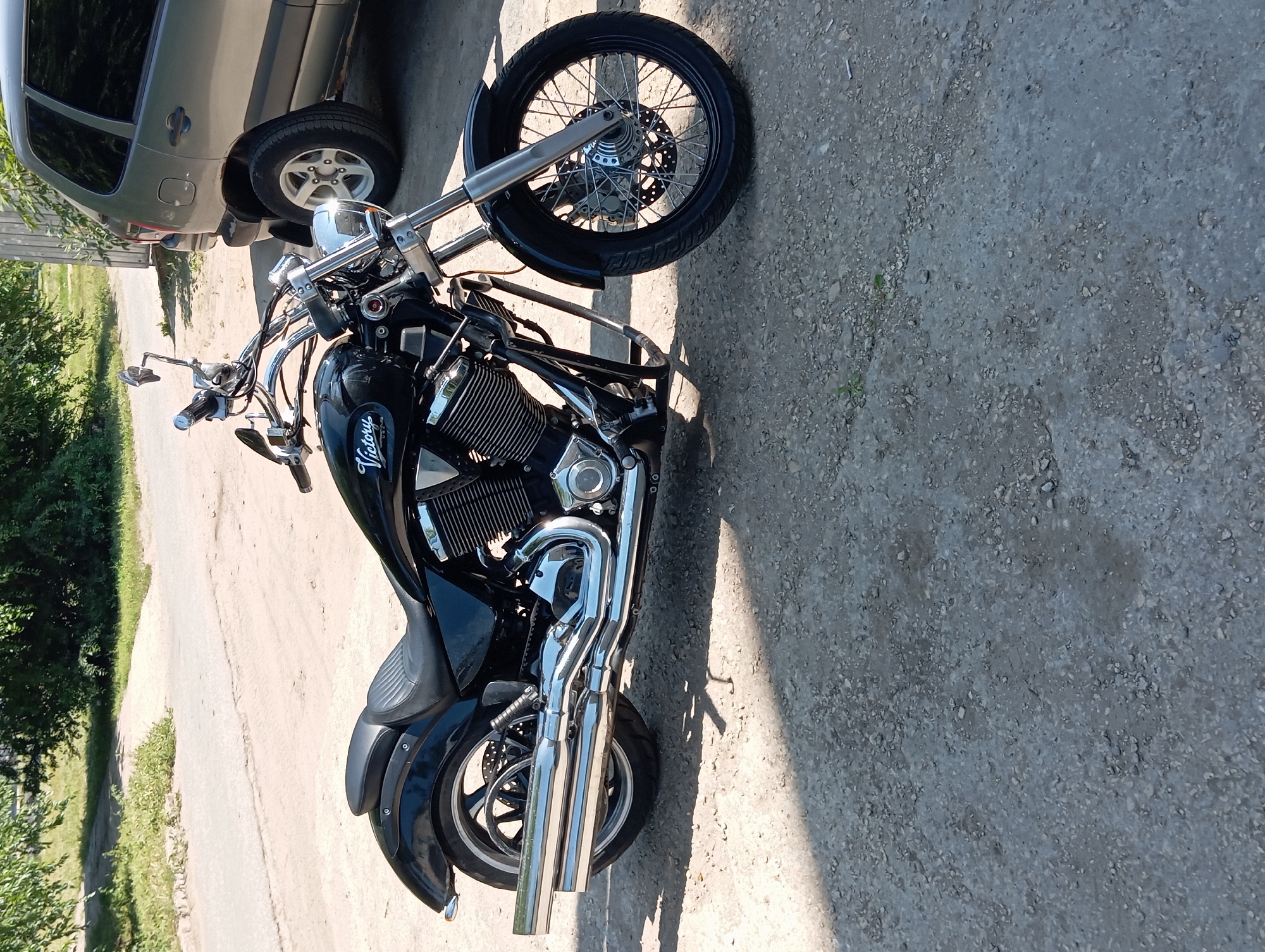 A black Victory motorcycle is parked on a gravelly surface in front of a silver car. The motorcycle is angled towards the right, with its front wheel turned slightly. The handlebars are chrome, and the engine is a V-twin. The motorcycle has dual exhaust pipes. The car is a hatchback and appears to be a older model. Behind the vehicles, there is a paved road with trees and foliage on the left side. The scene is outdoors, and the lighting suggests it is daytime. The image was taken in Balakovo, Russia.