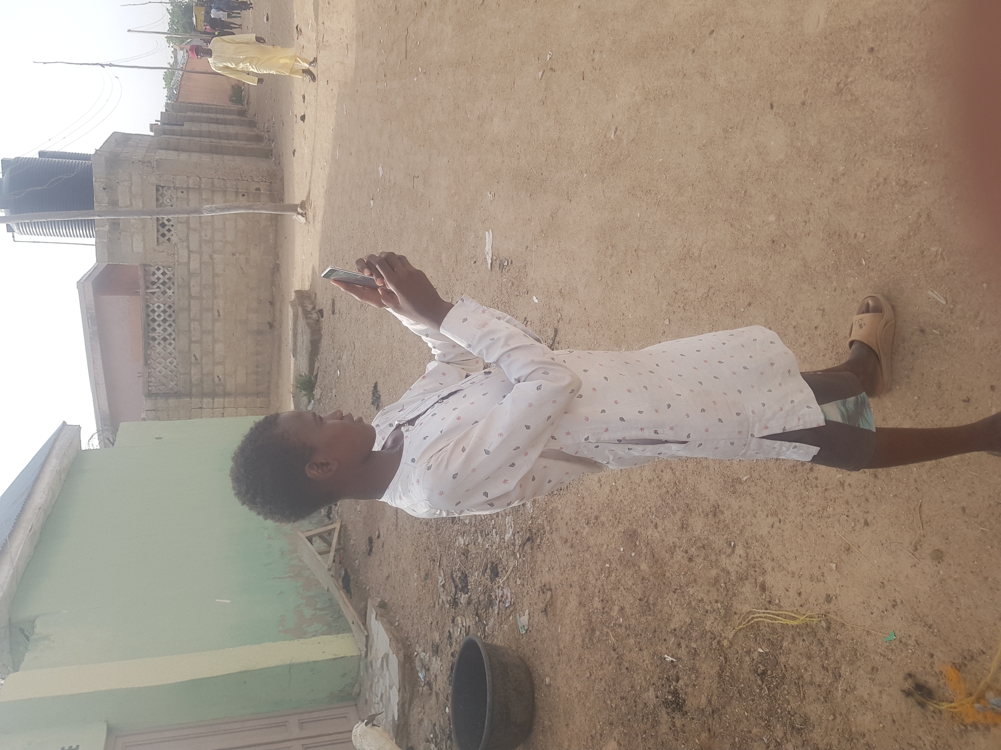 A young person in a white patterned shirt and sandals stands on a sandy ground surface, holding a smartphone in both hands, with their gaze directed at the device. A black bucket sits nearby. A green building is visible to the left, and a brick building and water tower are in the background. In the distance, a person in yellow clothing walks on the street in the direction of Dabawa, Nigeria.