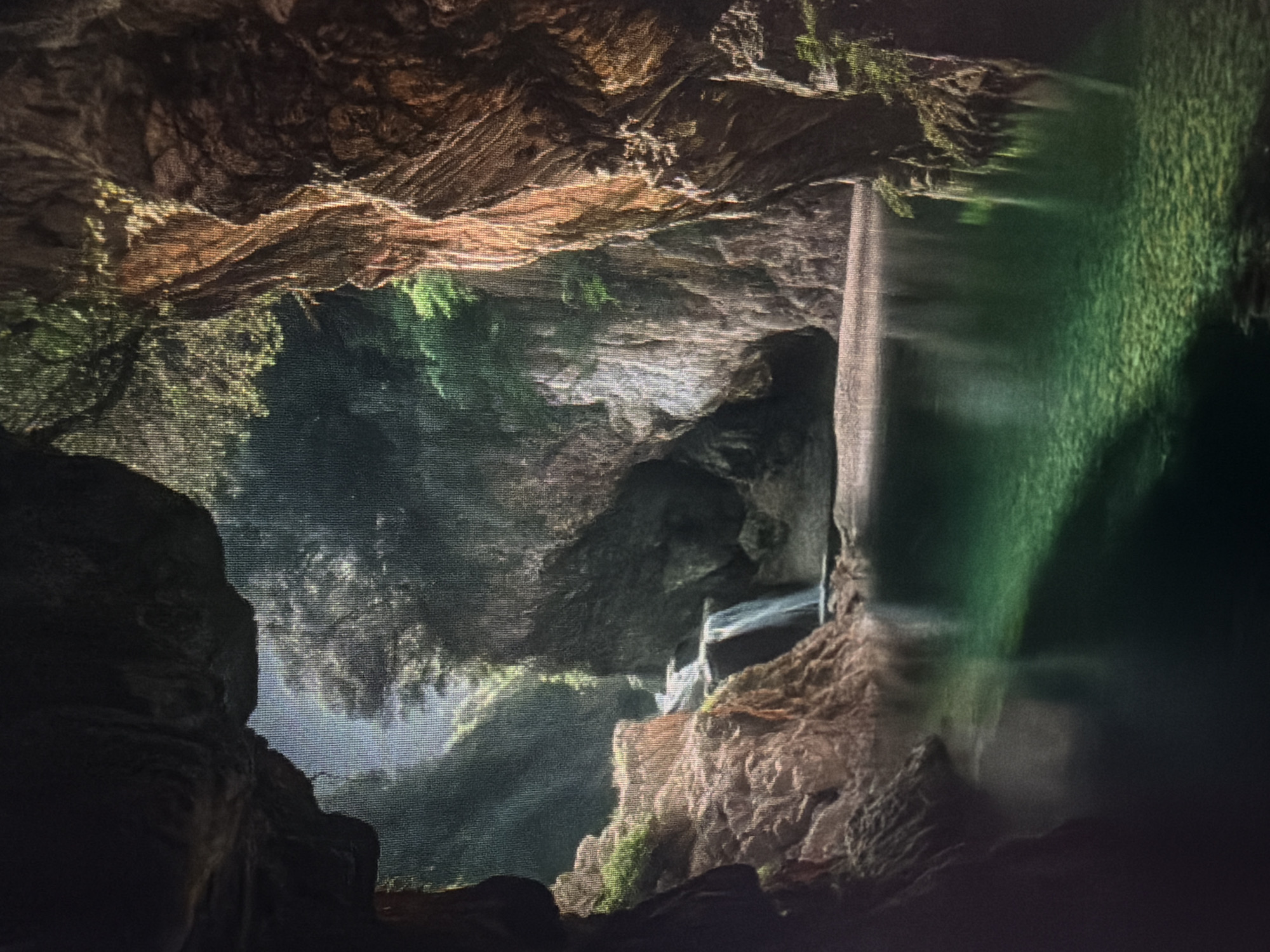 The image depicts a serene natural scene within a cave or grotto. A body of calm, clear, emerald-green water fills the foreground, with the rocky, textured bottom visible beneath the surface in the illuminated areas. To the left, a rocky outcrop with patches of green vegetation borders the water. Further into the scene, a small waterfall cascades down a rocky incline. Tall, rugged rock formations, covered in places with green foliage, flank the waterfall and the water body, creating a sense of depth and enclosure. Sunlight filters in from the opening, illuminating the scene and casting a soft glow on the water and rocks. The overall impression is one of a hidden, tranquil oasis. The location is identified as being in Baan Mae Nam, Thailand, suggesting a natural landscape within that region. There are no people or animals visible in the image.