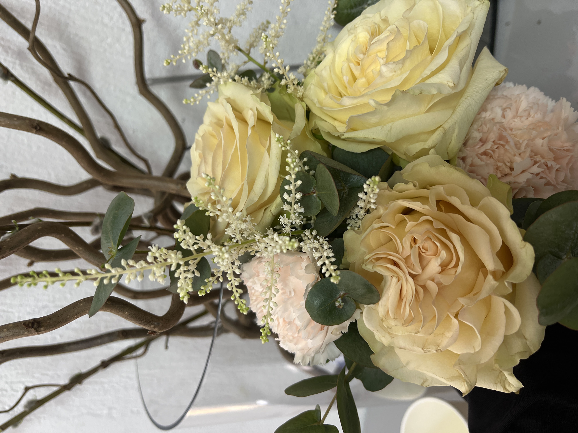 A still-life photograph presents a floral arrangement predominantly featuring roses and carnations, accompanied by eucalyptus leaves and delicate white flowering sprigs. The roses are a pale yellow-cream color, while the carnations are a subtle pinkish-peach. The arrangement is interspersed with eucalyptus leaves, small round leaves, and wispy white flowers. The backdrop consists of winding, bare branches, which add a rustic and textural element to the composition. The background is a plain white surface, and a portion of a white cup can be seen near the bottom right of the frame. The context and setting of Chaikovsky, Russia, are not evident from the visual information provided.