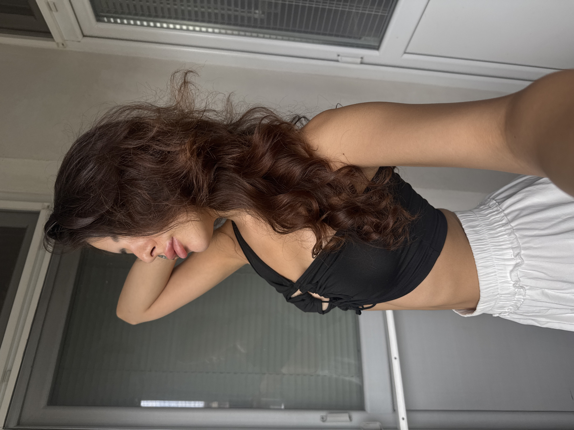 A young woman with dark, wavy hair is shown in profile, looking out a window. She is wearing a black crop top and white, high-waisted pants. Her left arm is raised, resting behind her head. The background features a closed window with blinds and a white door frame. The scene appears to be indoors, possibly on a balcony or in a room with a view of the exterior. The image does not provide any specific indicators to determine its location within the city of Plovdiv, Bulgaria.