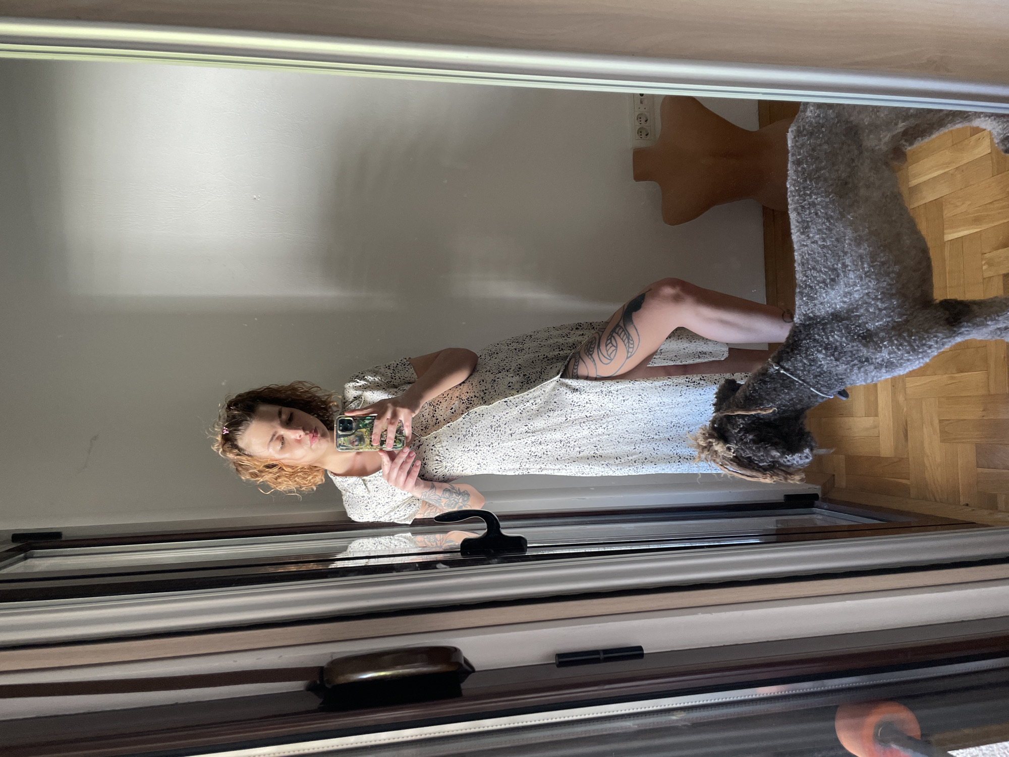 A woman with curly blonde hair stands in front of a mirror, holding a smartphone to take a selfie. She wears a light-colored dress with dark speckles, and tattoos are visible on her left arm and leg. Behind her, a grey dog stands on a wooden floor, near a dressmaker's dummy. The mirror is mounted on a window frame. The location is an interior space in Boreti, Montenegro. An electrical outlet is visible on the wall behind the dummy.