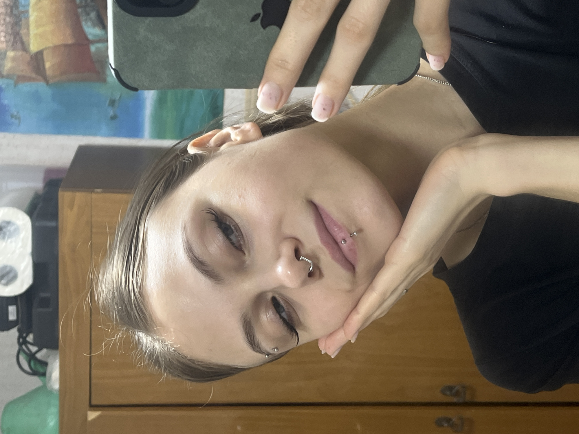 A person is shown in a selfie, holding a phone with a green case. They have fair skin, light brown hair slicked back, and piercings in their nose, lip, and eyebrow. The person wears a black shirt. The background includes a wooden dresser, a roll of paper towels on a shelf, and a blurred painting. There is no information in this image to identify the location as Фоки, Russia.