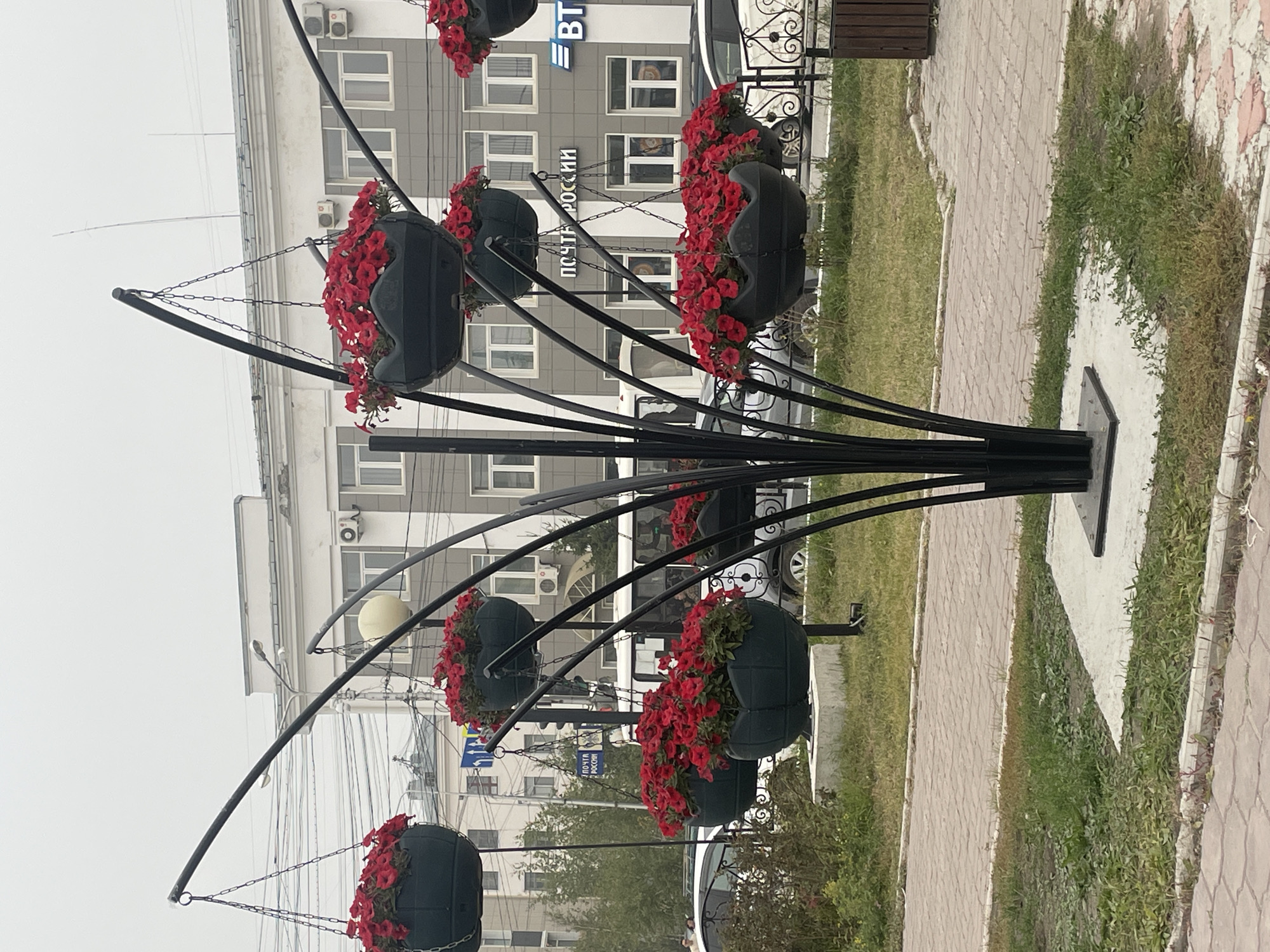 The image shows a decorative floral arrangement in the city of Yakutsk, Russia. The arrangement consists of multiple dark green hanging planters filled with red flowers, suspended from a black metal structure that resembles a stylized tree. The planters are strategically placed at varying heights. In the background, a modern building with multiple windows and air conditioning units is visible. A sign on the building reads "ПОЧТА РОССИИ" (Russian Post) and another logo, possibly for a bank, is also present. Several vehicles, including a white bus and cars, are visible on the street behind the floral display. The ground in front of the building is paved with brick-like tiles and bordered by grass and small plants. The sky is overcast, suggesting a cloudy day. There are no people clearly visible in the immediate foreground.