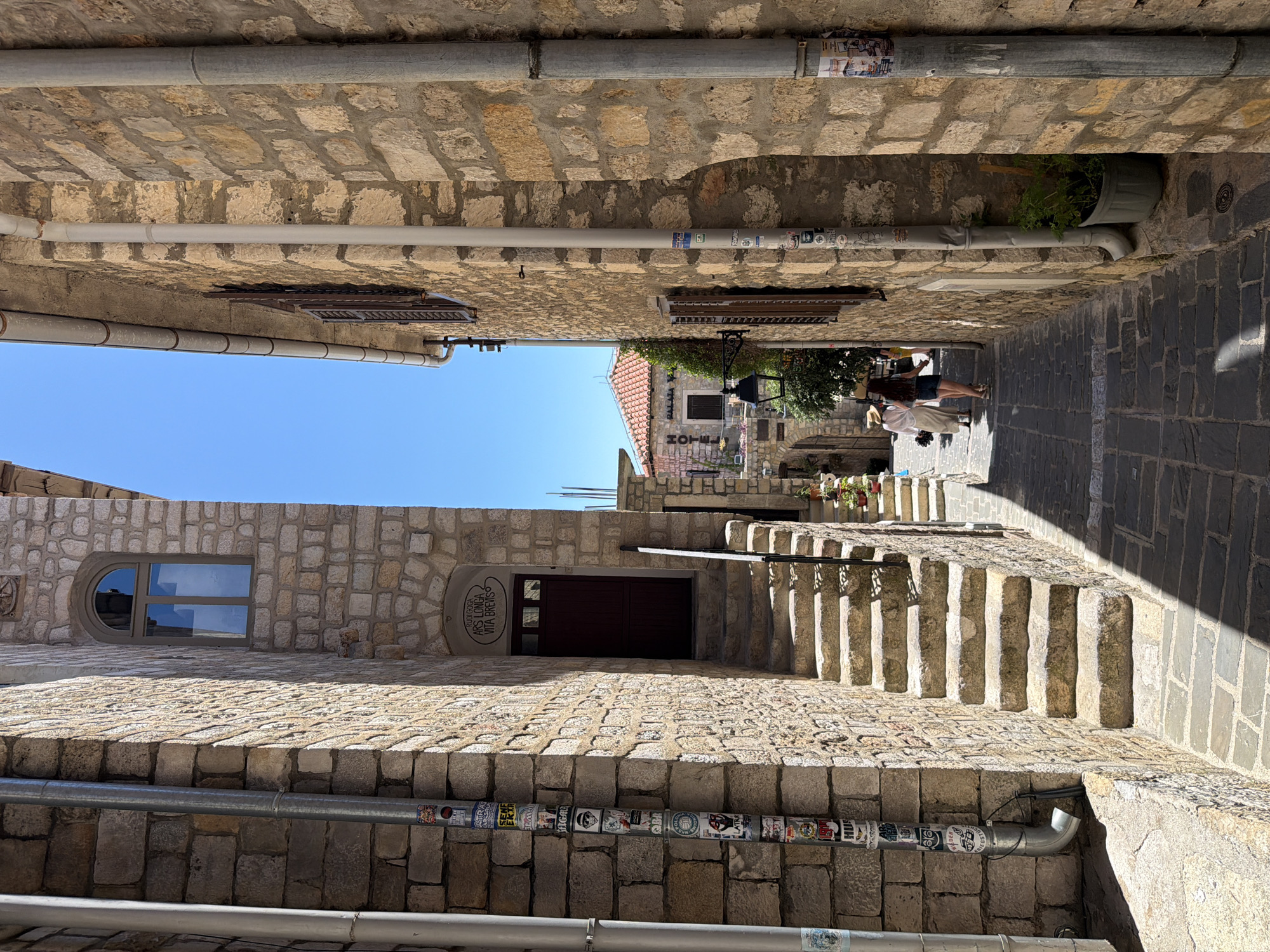 The image depicts a narrow cobblestone alleyway in the old town of Ulcinj, Montenegro. The buildings flanking the alley are constructed from rough-hewn stone, with one side featuring a tall, imposing stone wall adorned with numerous stickers on a large downspout. On the opposite side, a stone building has a doorway with a dark wooden door, accessed by a set of stone stairs. Above the doorway, a sign reads "RUDI GOGA ARS LONGA VITA BREVIS." Further down the alley, on the right, another stone building shows a window with dark shutters and a decorative lantern hanging outside. In the distance, a red-tiled roof is visible. Two individuals are walking away from the camera down the alley; one is wearing a white robe and a hat, and the other is a young woman in a denim skirt and a dark top. The sky above is clear and blue.