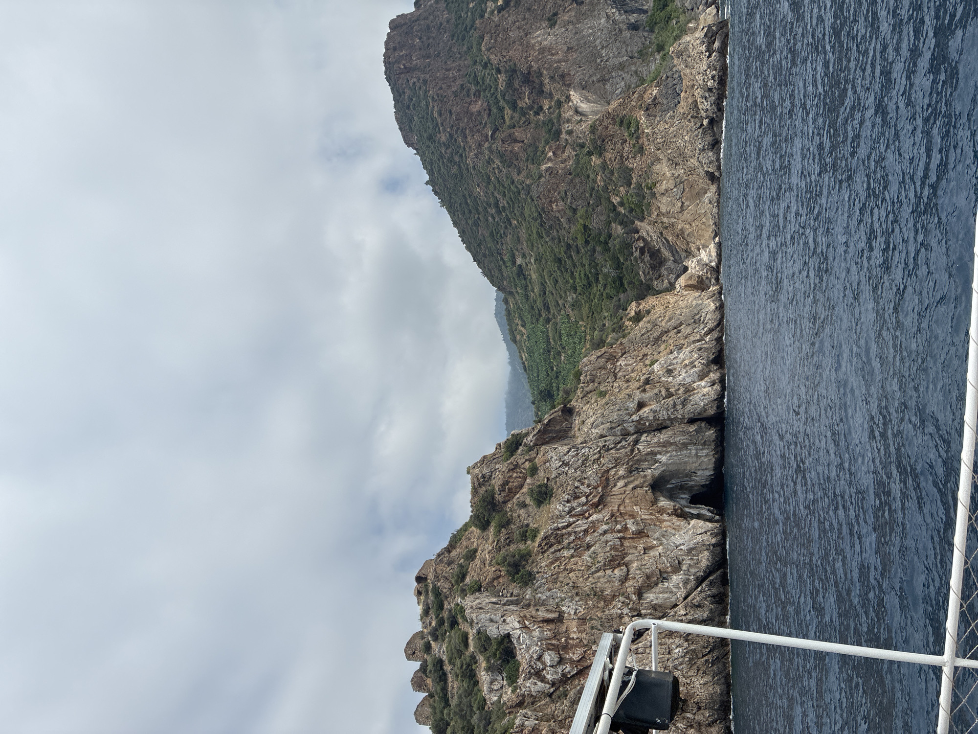 An eye-level, wide shot captures a rugged coastal landscape in Gazipaşa, Turkey, under an overcast sky. In the foreground, a white railing and part of a boat are visible, suggesting the vantage point is from a vessel on the sea. The dark, choppy water fills the lower portion of the frame.
Dominating the middle ground and background are imposing, rocky cliffs and hills. On the left, a steep, brown and gray cliff face features a prominent cave entrance at its base, its dark opening contrasting with the lighter rock. Above the cave, the cliff is sparsely vegetated with low green shrubs and scattered trees. Further up the slope, ancient-looking stone structures, possibly ruins, are visible on the crest of the hill.
To the right, a larger, darker mountain rises, its slopes covered in dense green vegetation. Terraced rows of what appear to be banana trees are visible on the mid-slope of this mountain. The distant horizon is obscured by more hazy, green hills. The overall scene conveys a sense of natural beauty and historical presence.