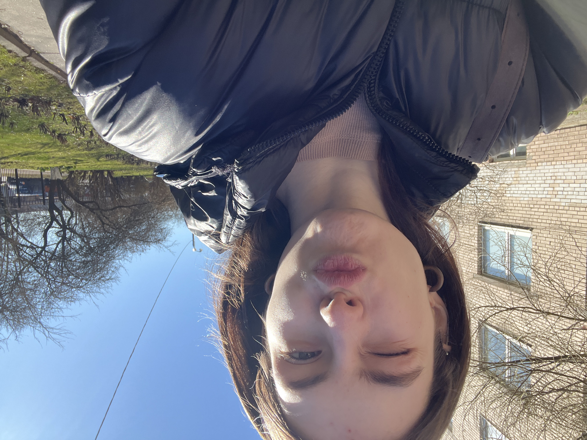A young woman takes a selfie outdoors. She wears a dark puffy jacket over a light pink top. Her expression is a slight pucker with one eye closed. The background is a clear blue sky, trees without leaves, a brick building with visible windows, and a strip of greenery along a road. This photo was taken in Saint Petersburg, Russia.