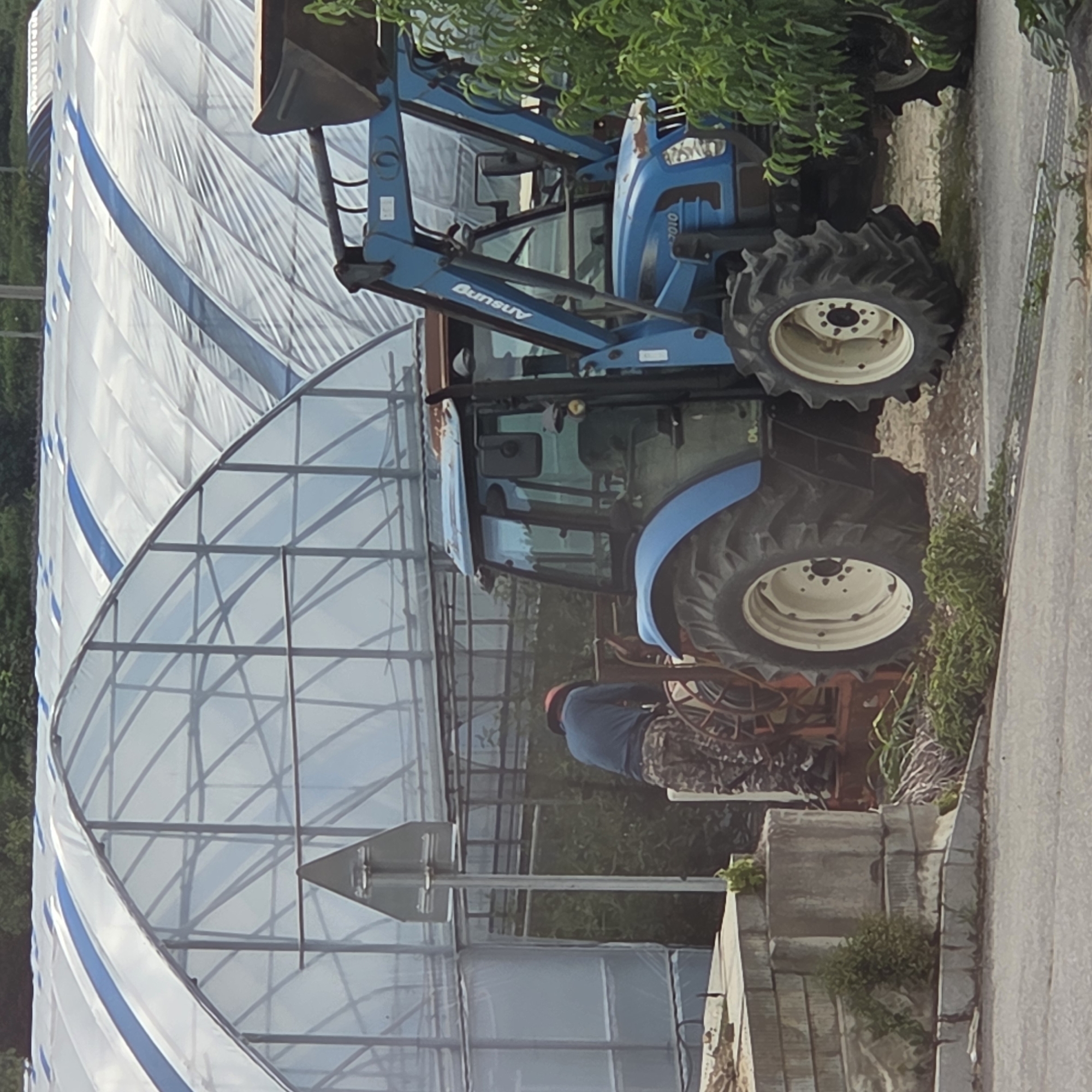 In Dunnae-myeon, South Korea, a blue tractor with a front loader is parked in front of a row of greenhouses covered with white plastic sheeting. A person wearing a blue long-sleeved shirt and camouflage pants is standing next to a piece of agricultural equipment, possibly a sprayer, attached to the back of the tractor. The tractor has the model number "2010" visible on its front. The environment is outdoors, with greenery visible around the tractor and in the background. A triangular road sign is also present, mounted on a metal pole.