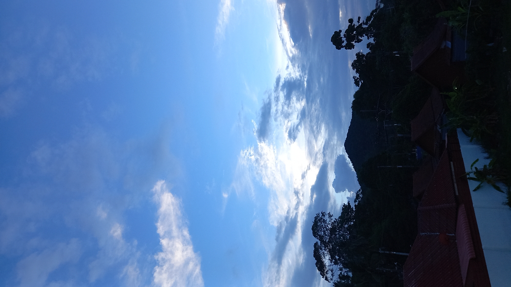 The image depicts a twilight scene in the Ko Pha-ngan District of Thailand, characterized by a dramatic sky and silhouetted landscape. The sky is a gradient of deep blue to lighter hues, with scattered white and grey clouds creating a textured ceiling. Sunlight is faintly visible breaking through the clouds in the upper right quadrant, suggesting the sun is either rising or setting.
Below the sky, a dark, densely forested hillside rises, with the silhouette of a prominent mountain peak visible in the center. The trees on the hillside are tall and appear lush, obscuring much of the terrain. Several power lines crisscross the mid-ground, supported by utility poles, indicating a populated or developing area within the otherwise natural landscape.
In the foreground, the rooftops of several houses are visible. These structures have characteristic red-tiled roofs, some with visible gables. A white wall runs along the bottom left of the frame, partially separating the residential area from the foreground vegetation. Small banana plants are interspersed amongst the houses, hinting at the tropical climate. The overall impression is one of a rural or semi-rural residential area nestled against a backdrop of natural, mountainous terrain under a dynamic sky. No people are visible in the image.
