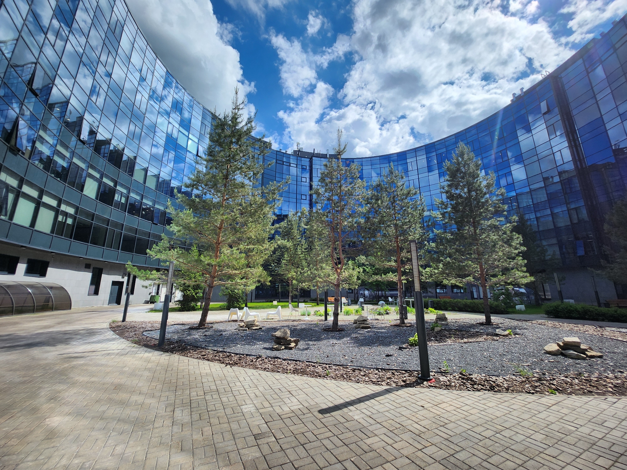 The image shows a courtyard surrounded by a curved modern office building in Unknown, Unknown. The building has a glass facade reflecting the sky and clouds. In the center of the courtyard, there is a circular area filled with dark gravel mulch and several tall pine trees. Between the trees, there are visible benches and chairs. The ground is paved with interlocking bricks. Modern lamp posts are placed around the tree bed.