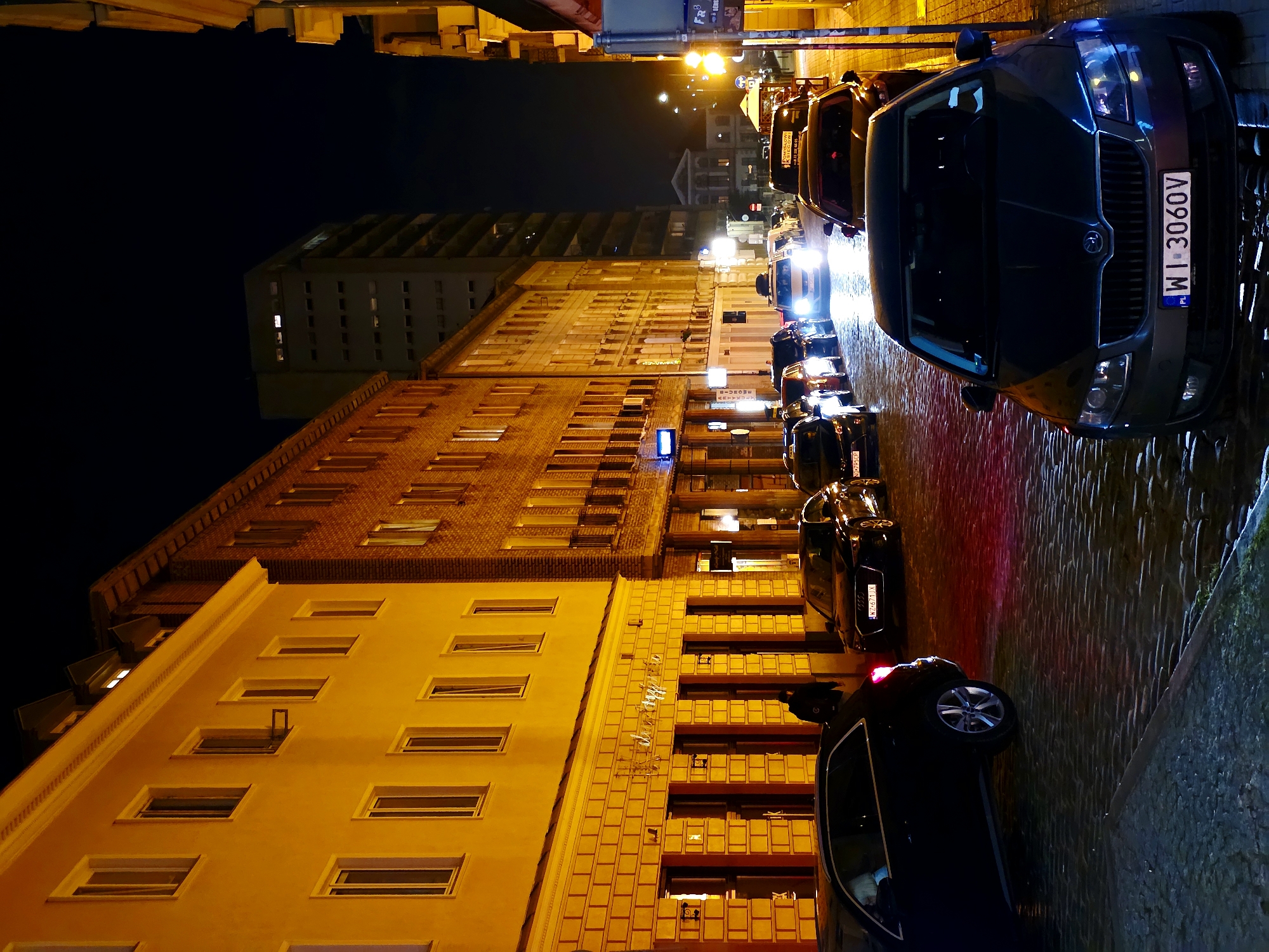 The image depicts a nighttime street scene in Warsaw, Poland. The street is paved with cobblestones, and buildings line both sides of the street. The buildings are illuminated by warm yellow and orange lights, creating a cozy ambiance. Several cars are parked along the street, including a black Audi with a license plate reading "WZ 671 XX" and a dark grey Skoda with a license plate reading "WI 3060V". A person in a dark jacket is walking away from the camera on the sidewalk. In the background, a tall, modern building stands out against the dark night sky. The overall scene suggests a vibrant city nightlife, with illuminated streets and cars hinting at the ongoing activities in Warsaw.