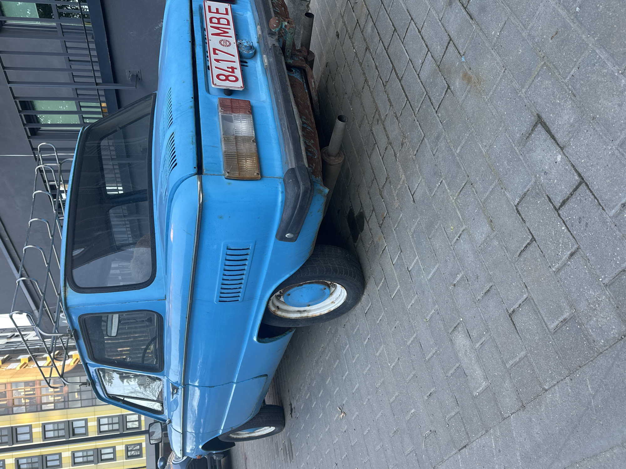 The image shows a blue ZAZ-968 parked on a grey brick sidewalk. The car has a roof rack and a license plate reading "8417 MBE". The car appears to be in a state of disrepair, with visible rust. The sidewalk is made of rectangular grey bricks laid in a running bond pattern. In the background are buildings with black and white facades. The image was taken in Minsk, Belarus.