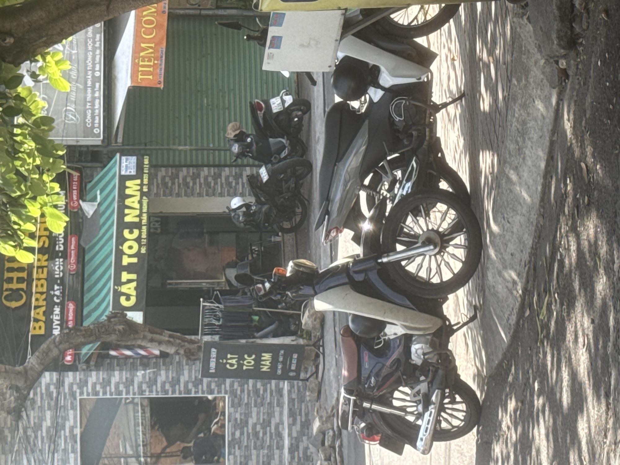 In Bac Nha Trang Ward, Vietnam, a street scene unfolds with a barber shop prominently featured. A sign above the entrance reads "CHI BARBER SHOP" with "HUYEN: CAT-UON-DUOI" below, indicating hair cutting, perming, and styling services. Further down, another sign declares "CAT TOC NAM" (Men's Haircut) with the address "DC: 12 DOAN TRAN NGHIEP" and phone number "DT: 0935 812 683". Adjacent to this, a vibrant green awning shades the facade. To the right, a sign in orange text announces "TIEM COM" (Rice Restaurant).
Several motorcycles are parked along the sidewalk. In the foreground, a dark blue and silver motorcycle is positioned on the left, facing right, with its kickstand down. To its right, a grey and white motorcycle is parked, also facing right. Further back, two other dark motorcycles are visible. A tall, slender tree with sparse foliage stands to the left of the barber shop, partially obscuring it and the barber pole. The ground is paved with light-colored bricks, with shadows from the tree and possibly awnings falling across it. A stone curb runs along the edge of the sidewalk. The overall impression is a typical commercial street in a Vietnamese city.
