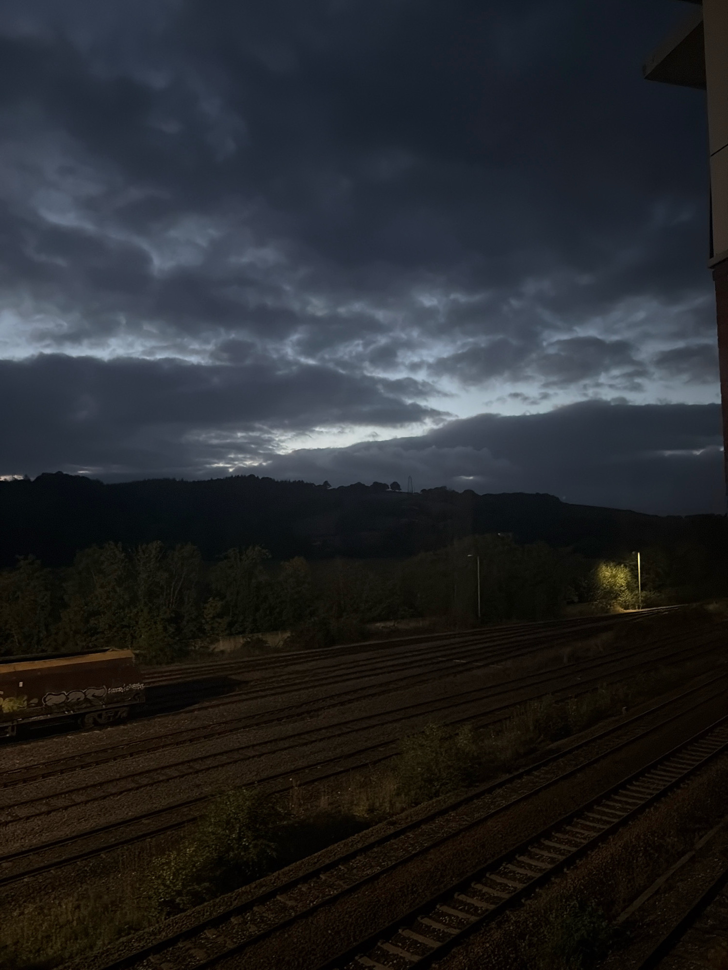 The image depicts a twilight scene of a train yard with multiple parallel railway tracks stretching into the distance. A brown train car, marked with graffiti, is parked on the leftmost tracks. Beyond the tracks, a dense line of trees forms a dark silhouette against a hill covered in vegetation. The sky is filled with dramatic, dark, and cloudy formations, with a sliver of lighter sky visible near the horizon. Two streetlights are visible in the mid-ground, one casting a bright pool of light on the trees and ground. The scene is captured from an elevated perspective, possibly from a building, as indicated by a sliver of a building edge on the right side of the frame. The overall atmosphere is moody and somewhat ominous, suggesting approaching night or stormy weather. The location is identified as Exeter, United Kingdom.