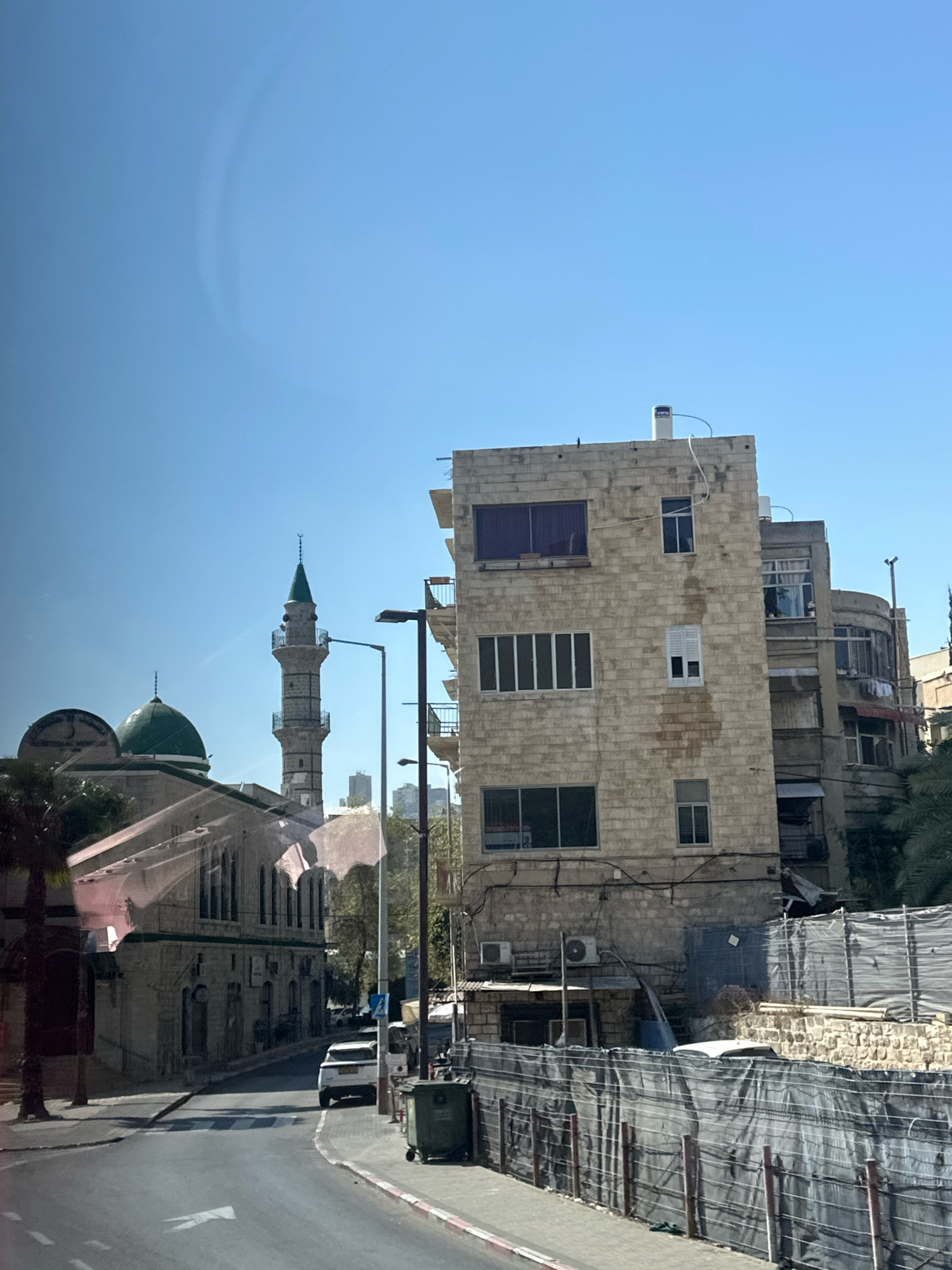 The image captures a street scene in Haifa, Israel, featuring a blend of architectural styles under a clear blue sky. On the left, a mosque with a green dome and a tall, slender minaret dominates the skyline. The minaret is made of stone with decorative bands and a pointed, finial-topped roof. Adjacent to the mosque is another building, possibly part of the same complex, with arched windows.
To the right of the mosque, a multi-story building constructed from light-colored brick stands prominently. It has numerous windows, some with dark glass and others with white shutters. There are visible external pipes and air conditioning units attached to the building's facade. Further to the right, other buildings with balconies and varying architectural details are visible, including a curved facade on one of them.
The street in the foreground is paved with asphalt and marked with a white arrow indicating direction. A white car is parked on the side of the road, and a green dumpster is situated on the sidewalk. A fence made of dark mesh and wooden posts runs along the right side of the street, separating it from a construction area or undeveloped land behind it, which is partially covered by the mesh and reveals stone walls. Palm trees and other greenery are visible, suggesting a Mediterranean climate. The overall impression is an urban environment with a mix of historical religious structures and modern residential buildings.