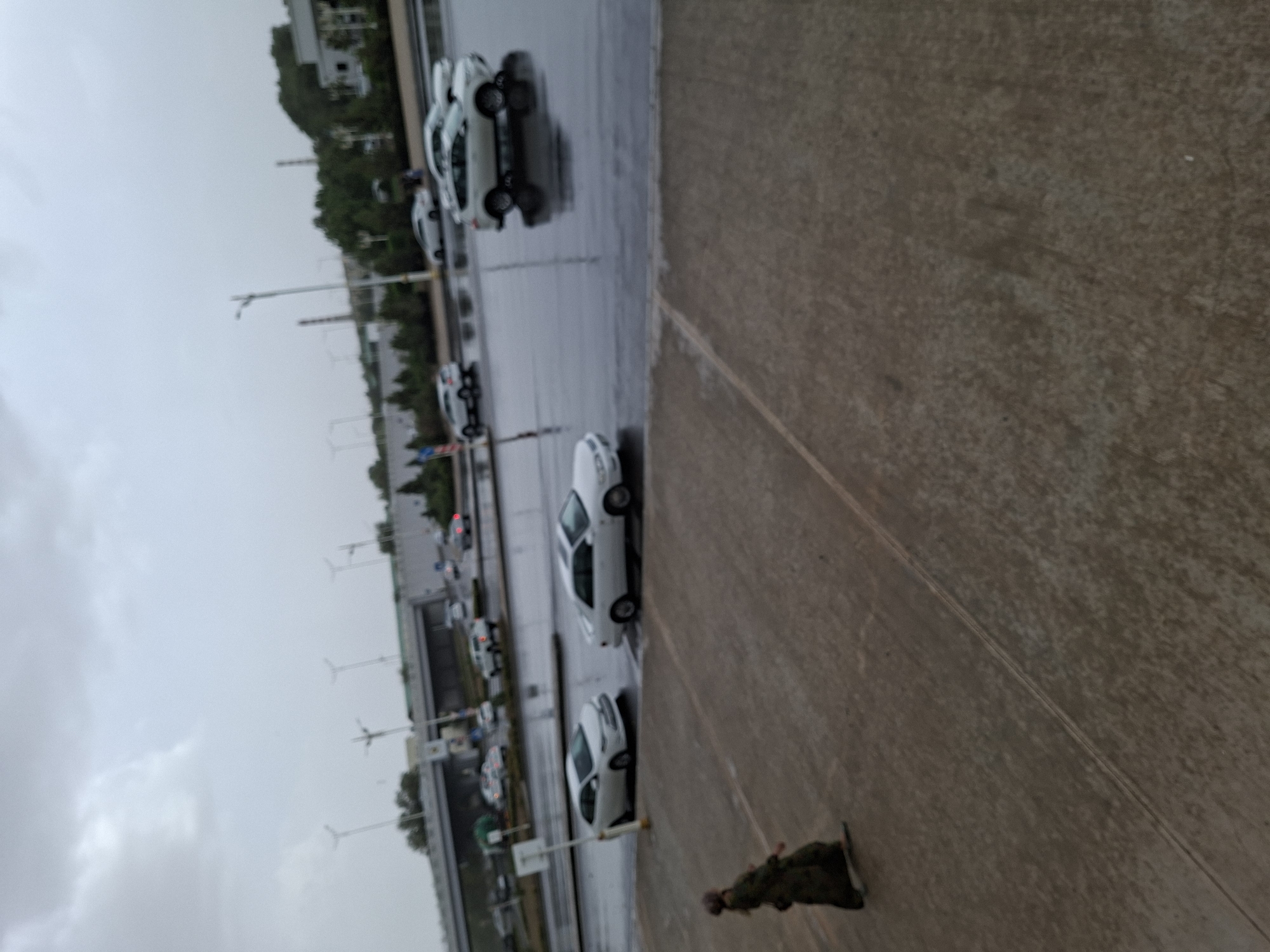 The image depicts a street scene in Ashgabat, Turkmenistan, under overcast and likely rainy conditions, suggested by the wet asphalt and reflections. In the foreground, a person wearing a dark, long garment and a headscarf walks across a paved area. Beyond this, a road is visible with several cars. Two white sedans are prominently featured on the wet road, one appearing to be parked or stopped, and the other in motion. Further in the background, more vehicles are present on the multi-lane road, and a highway overpass structure can be seen. Trees and buildings are visible on the right side of the image, and streetlights are scattered throughout the scene. The overall impression is of an urban environment experiencing inclement weather.
