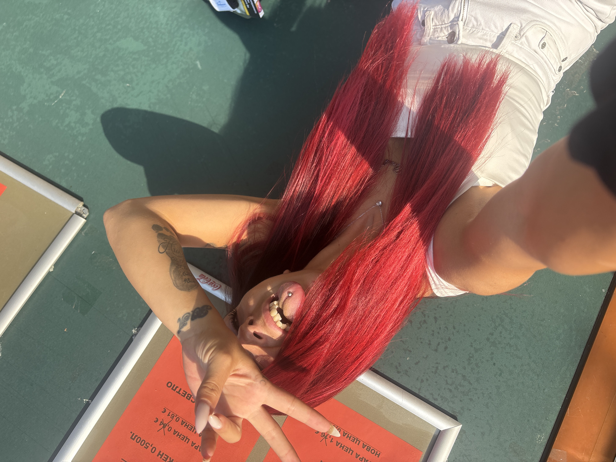 A young woman with bright red hair is lying on a dark green surface, possibly a bench or table, outdoors. She is wearing a white top and white pants. Her right hand is raised, with fingers extended and partially covering her face, and she is making a peace sign with her left hand. She has a tongue piercing with a silver ring and her tongue is slightly out. There are tattoos on her arm. A red sign with Cyrillic text is visible to her left. The scene appears to be in Sofia, Bulgaria, as indicated by the Cyrillic text on the sign. Sunlight casts a shadow on the surface.