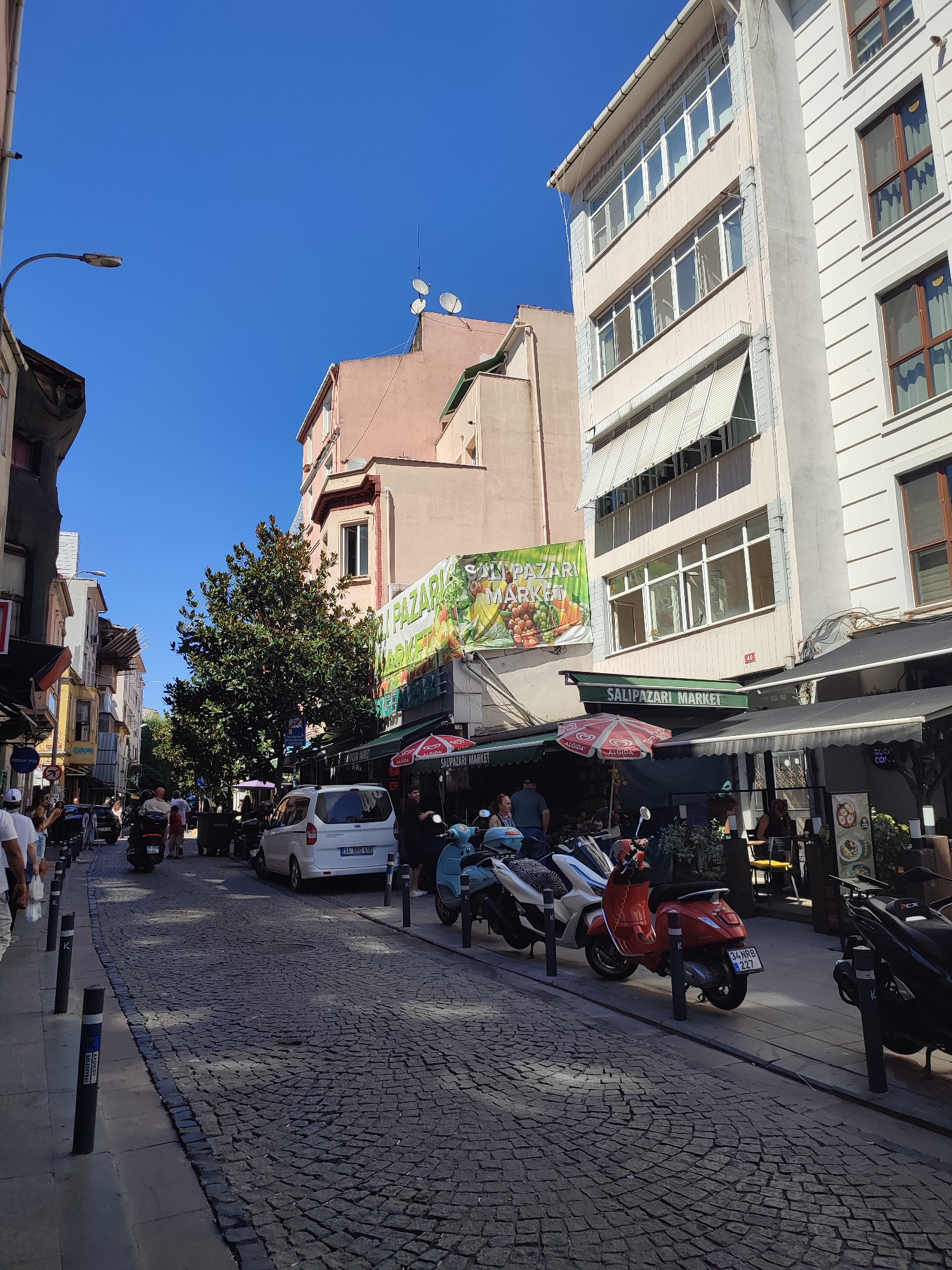A street in Kadıköy, Turkey, is depicted on a bright, sunny day under a clear blue sky. The street is paved with cobblestones and lined with buildings and market stalls. On the right side of the street, a modern, multi-story building with a light-colored facade and large windows stands tall. Below it, market stalls are set up with awnings, featuring a sign that reads "SALIPAZARI MARKET". Several red and white umbrellas are displayed at these stalls. Parked scooters and motorcycles are visible along the street. A white van is parked further down the street.
On the left side, older buildings with a more traditional architectural style are visible, some with shops and awnings. A street lamp with a curved arm is attached to one of these buildings. People are seen walking on the sidewalk and on the street, some are riding motorcycles. A large green tree stands in the middle of the street, between the market stalls and the buildings. A banner hangs on a building in the background depicting fruits and vegetables, also advertising "SALIPAZARI MARKET". The overall impression is of a bustling urban street scene with market activity.