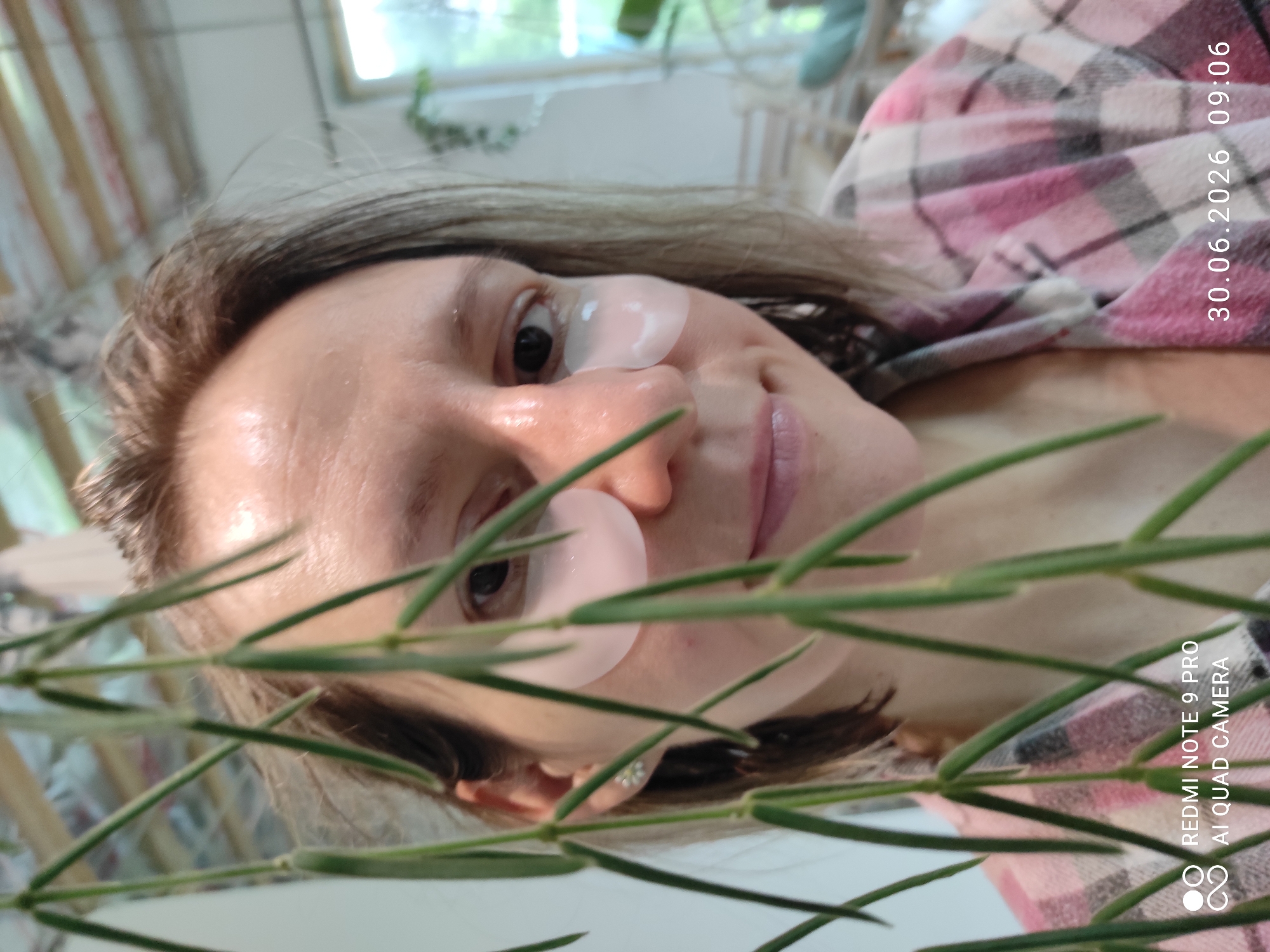 A person with light brown hair, wearing a pink and grey plaid shirt, is shown from the chest up, looking at the camera. The person has two pink under-eye patches applied. Green vines are obscuring the left side of the image. The background is blurred, with what appears to be a window and plants visible on the right. A timestamp in the lower right corner reads "30.06.2026 09:06". A watermark in the lower left corner indicates "REDMI NOTE 9 PRO AI QUAD CAMERA". The image does not provide any information about the city of Malyi Bukor, Russia.