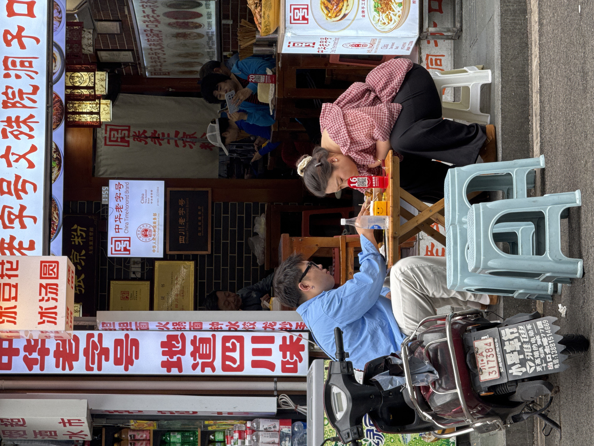 The image shows an outdoor scene in the Jinjiang District of Chengdu, China, with focus on a group of people sitting at a small wooden table, and a scooter parked nearby. A woman in a red and white checkered top is leaning over the table with her head down, looking at food, while a man in a light blue shirt and tan pants is sitting across from her, pouring a bottle of water. Behind the woman are two other people seated, one looking at a cell phone, and another with a white cap. Several signs, including a large vertical sign with red Chinese characters, advertise local food, and a China Time-honored Brand sign. A scooter is parked to the right, bearing a license plate that reads "317384."