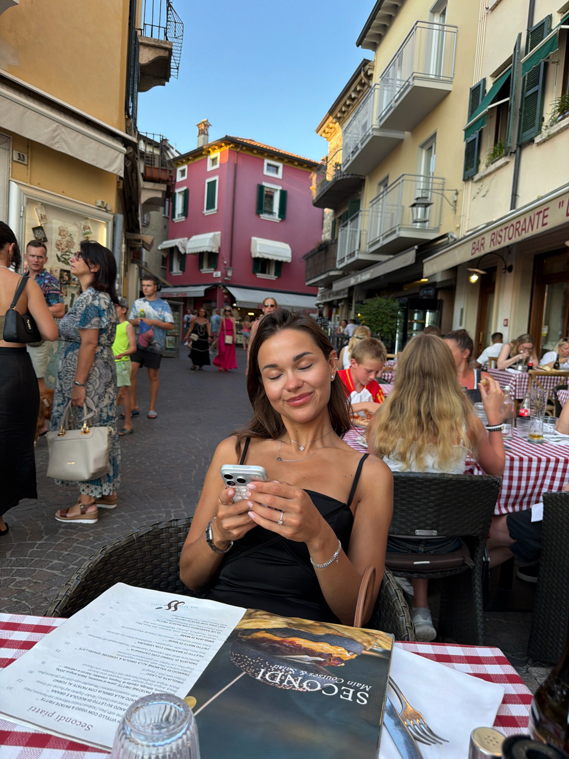 A woman is seated at an outdoor table in a narrow, cobbled street in Sirmione, Italy, looking at her phone. She is wearing a black dress and has a silver necklace and bracelets. A menu with a picture of a burger and the text "SECONDI Main Courses & SALADS" is laid out on the red and white checkered tablecloth in front of her, along with a glass and silverware. In the background, other diners are visible at tables, and the street is lined with colorful buildings with balconies and shops. The sky is clear and blue.