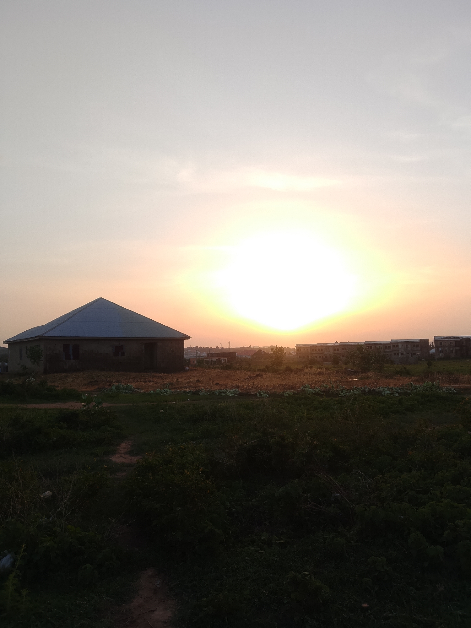 The image shows a rural scene at dusk or dawn in Ibo, Nigeria. A small, unpainted, single-story house with a corrugated metal roof stands in a field of low-lying vegetation. The sun is positioned low in the sky, creating a bright, hazy glow that obscures details in the middle distance. In the background, there are unfinished or partially constructed buildings. The foreground is dominated by dark green vegetation and a dirt path.