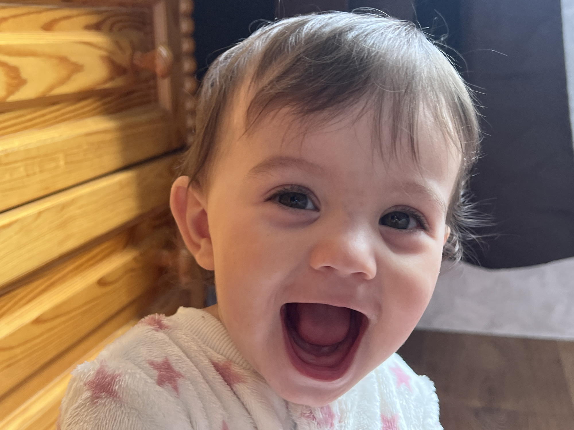 A close-up, slightly angled portrait shows a young child with light brown hair and fair skin. The child is wearing a white sweater with pink star patterns. The child's mouth is wide open in a smile or laugh, revealing their tongue. Behind the child, there is a wooden structure with horizontal planks on the left and a dark, solid surface on the right. The photograph appears to be taken indoors. There is no visual information to confirm its location in Elena, Bulgaria.