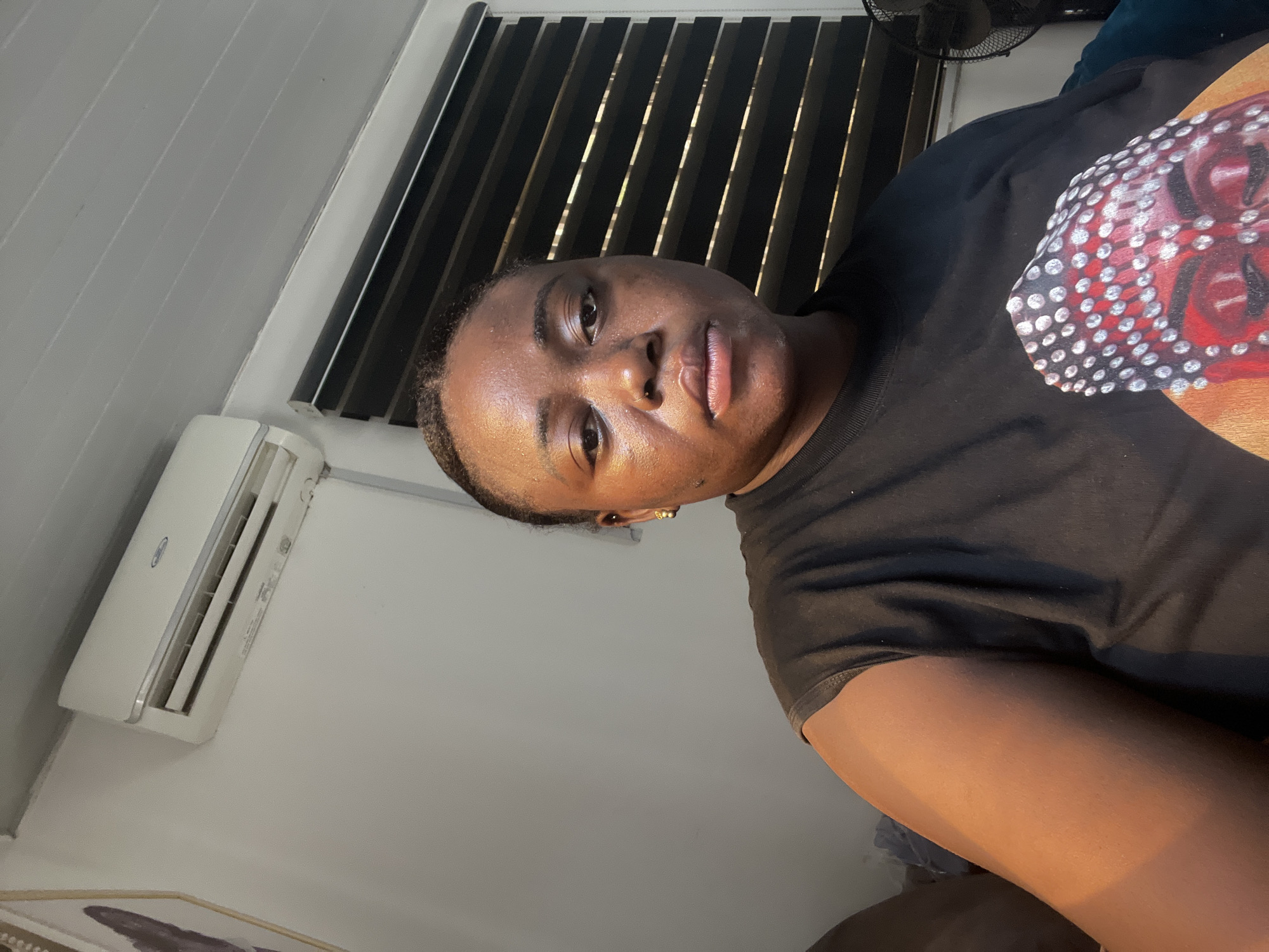 The image shows a person with dark skin wearing a black t-shirt. The person is indoors, near a window with closed blinds and an air conditioner unit. The location is likely within a building in Abuja, Nigeria. A fan is visible in the upper right corner. There is also a portion of a framed piece of art visible in the lower left corner. The person is looking directly at the camera. The t-shirt has a red and white design on the shoulder.
