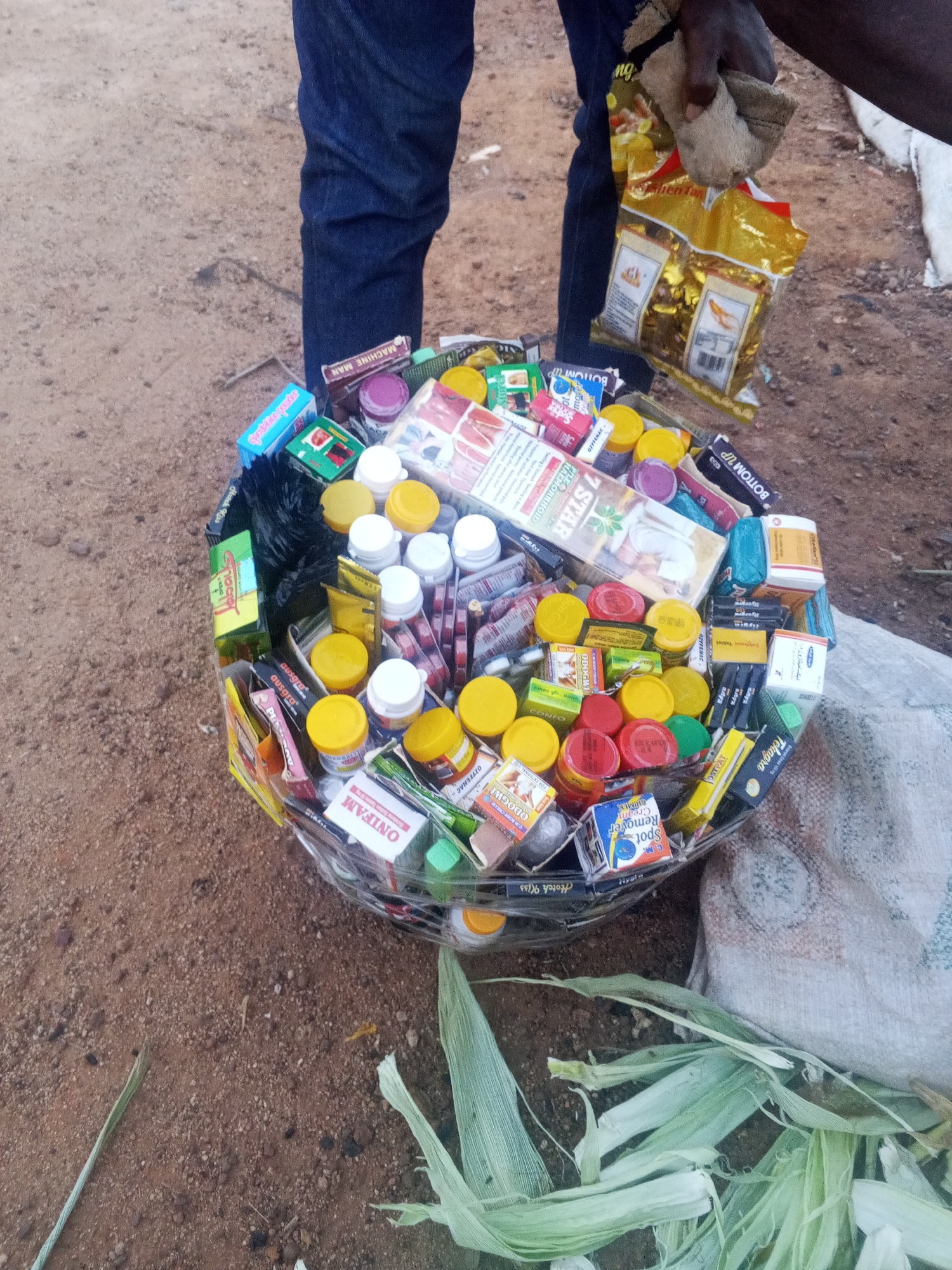 The image shows a person standing outdoors in what appears to be a dirt area. The person is holding a bag of "Men Tan". A large plastic container filled with numerous medicine boxes and jars is situated between the person's legs. The medicine boxes include the brands “7 STAR HAEMORRHOID”, "ONIFAM", "BOTTOM UP" , "CM", "DOGWU", "Hotch Kiss", "Ausgra", "Tehagra", "Zshifranil Tablet", "MACHINE MAN", "OZYFENAC", "OSTPAT", and "CONFO". Some corn leaves are scattered on the ground near the container. Based on the OCR content, the city is Bukuru, Nigeria.