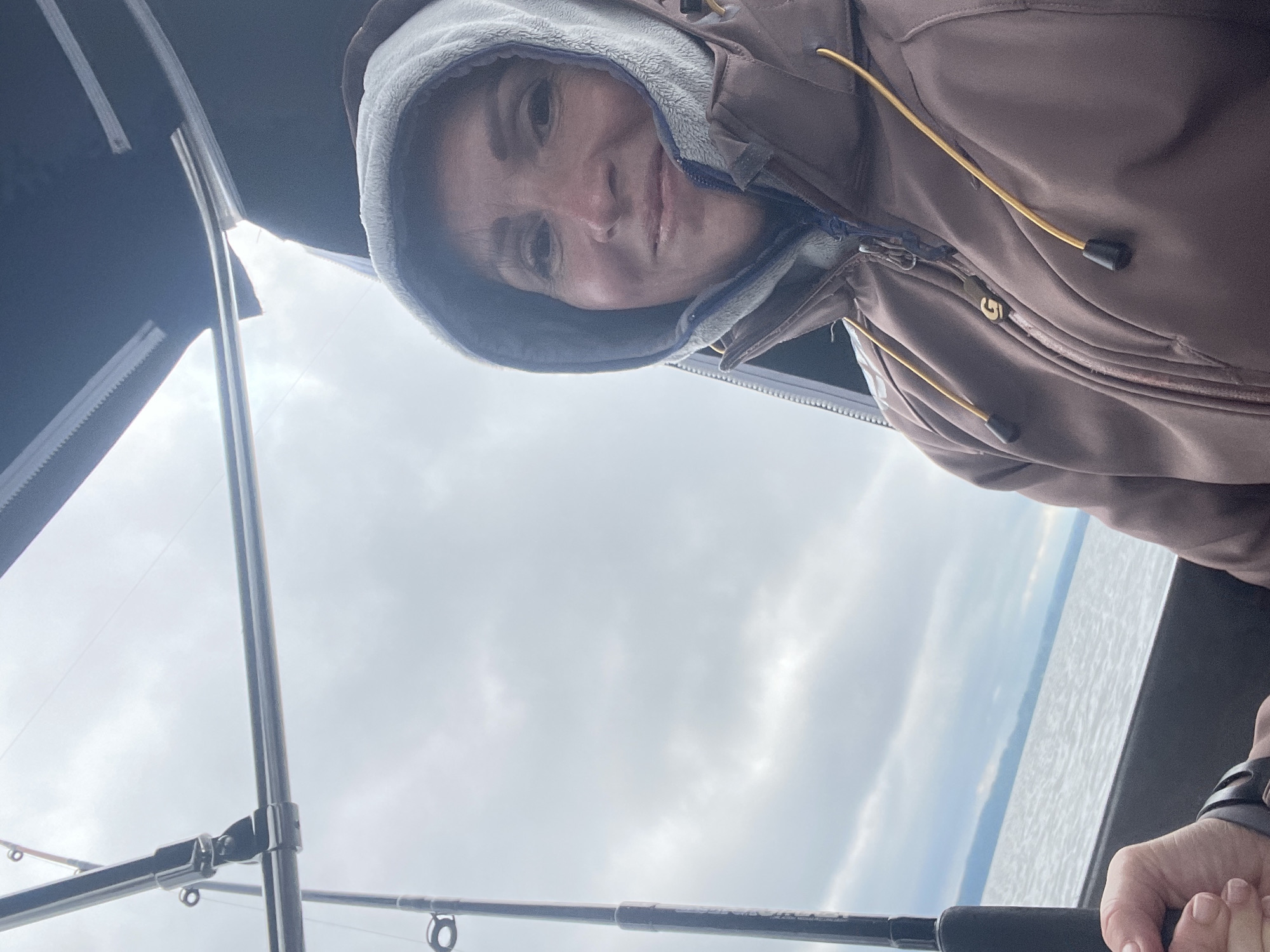 A woman wearing a hooded jacket and a grey inner layer smiles at the camera, facing right. She is on a boat, with fishing rods visible on the left side of the frame and a partial view of a canopy overhead. The water and a distant shoreline are visible in the background under a cloudy sky.
