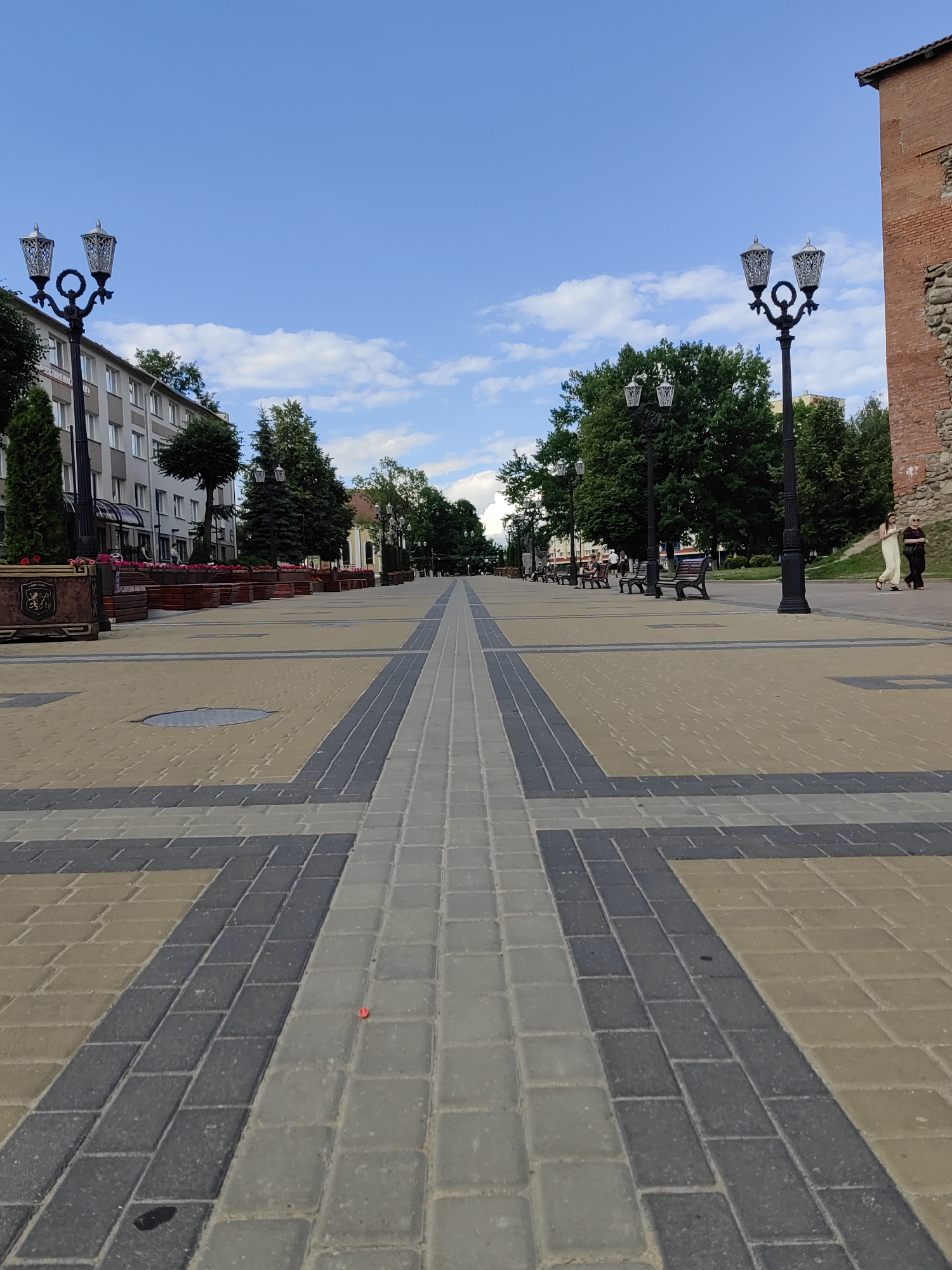 This is a wide shot of a pedestrian walkway on a sunny day, with a clear blue sky dotted with white clouds. The walkway is paved with interlocking rectangular stones in alternating patterns of yellow, gray, and dark gray. Tall, ornate black lampposts line both sides of the walkway, casting long shadows.
On the left side of the walkway, there is a multi-story building with a modern facade, featuring numerous windows and balconies. Planters filled with vibrant pink flowers and dark green foliage run along the base of the building. A few trees are interspersed along the walkway.
On the right side, beyond the lampposts and trees, a large brick structure with visible stone ruins is present, suggesting historical significance. A few people are visible walking on the far side of the walkway, some sitting on park benches under the shade of trees. The overall atmosphere is peaceful and leisurely, suggesting a public space enjoyed by locals and possibly tourists. The time of day appears to be late afternoon, given the length of the shadows. No discernable text is visible in the image.