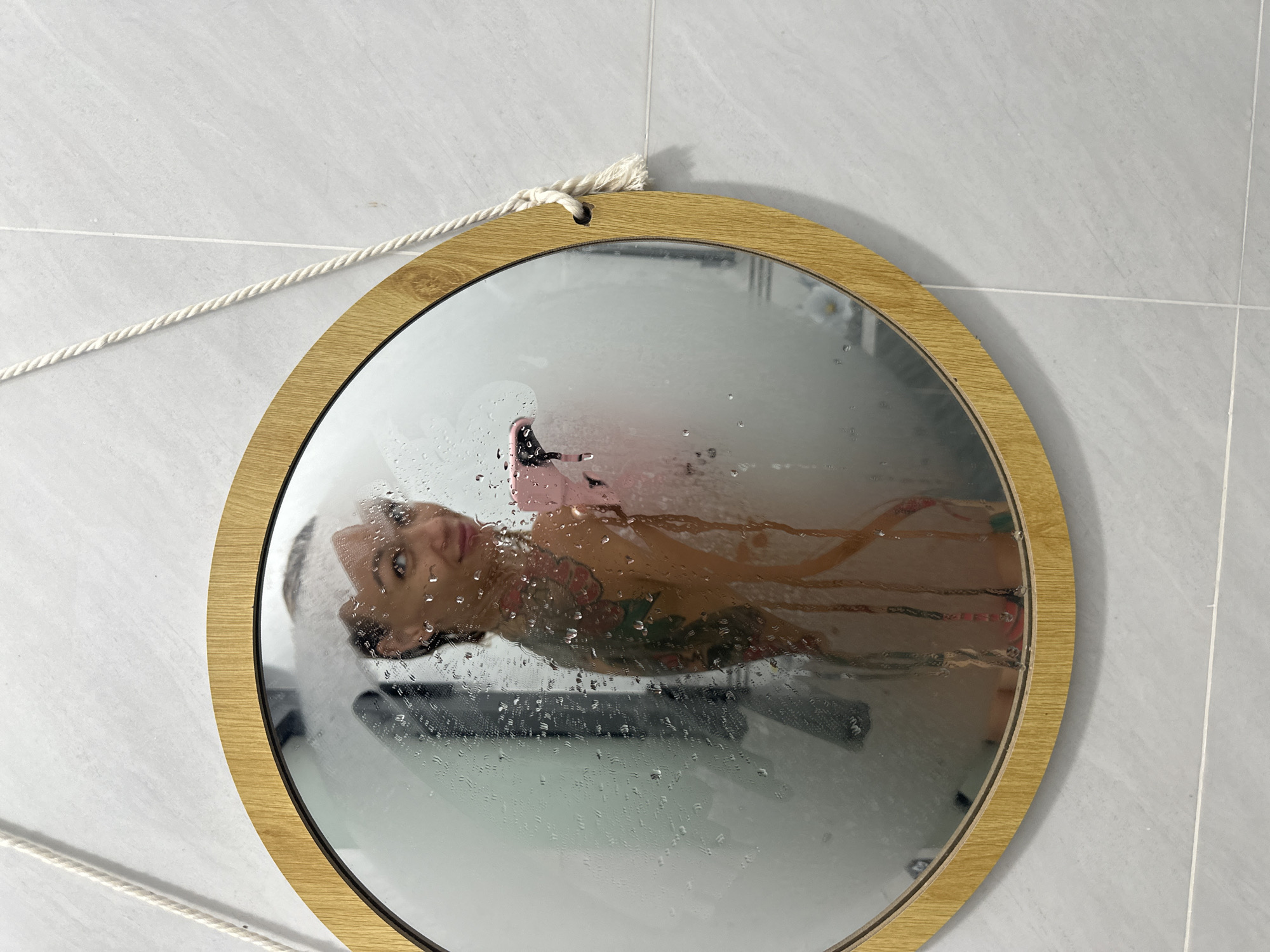 A woman is reflected in a steamed-up, oval mirror hanging by a rope against a tiled wall. The mirror has a light-colored wooden frame. The woman appears to be in a shower, holding a pink object, possibly a phone, and looking at the camera with a slight smile. She has tattoos on her arm and shoulder. The background appears to be a bathroom with light grey tiles. There is no indication of the city of Mũi Né, Vietnam.
