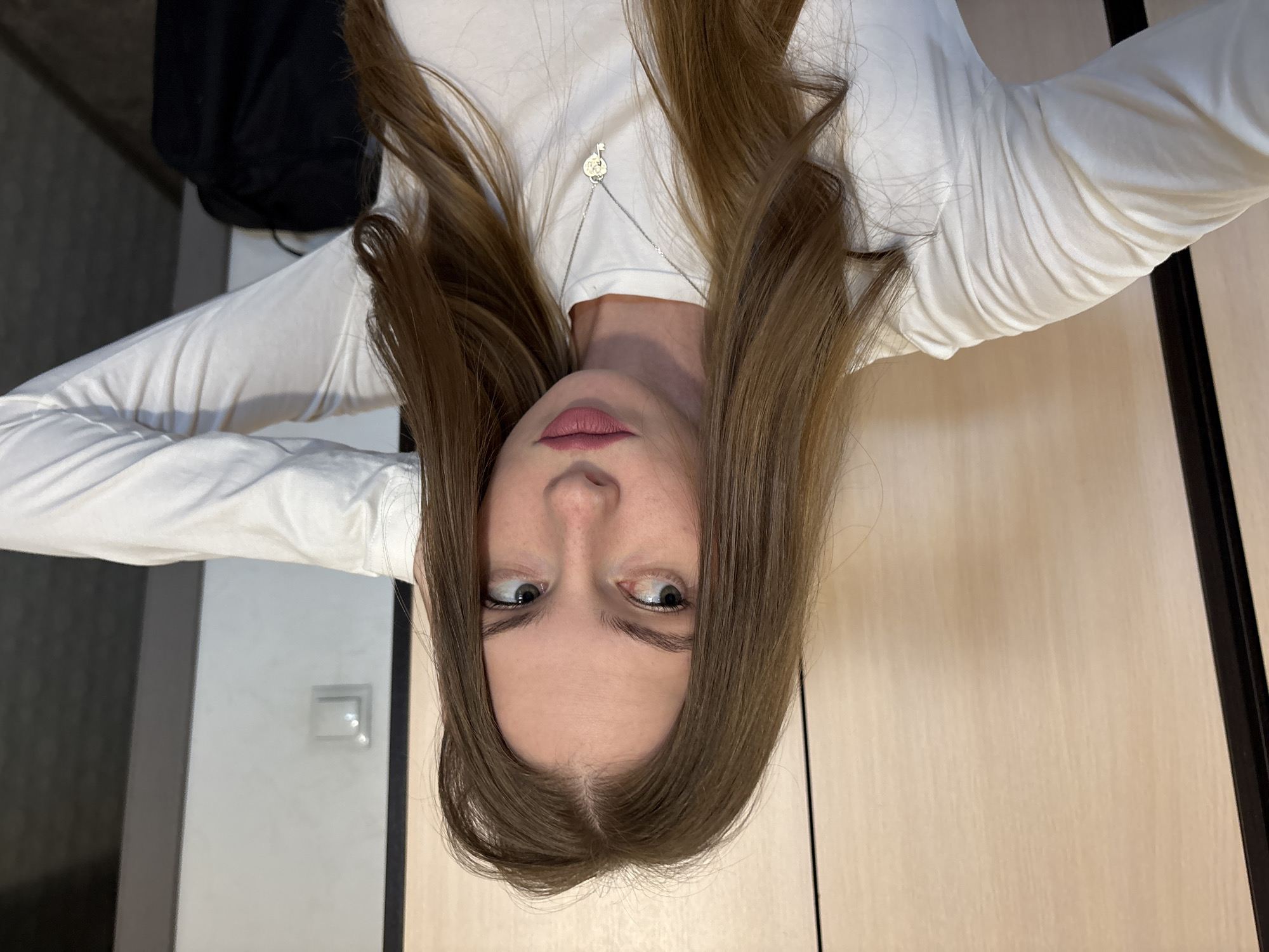 The image shows a young woman with long, light brown hair, wearing a white long-sleeved shirt and a silver necklace with a key pendant. She is looking to her left with a slight pout. Her right hand is raised and touching the back of her head, while her left arm is bent and extended forward. The background appears to be an indoor setting, possibly a closet or a room with wooden-paneled walls. A light switch is visible on the wall to her right. The scene is lit with natural light, casting soft shadows. There is no indication of the city of Perm, Russia, in the image.