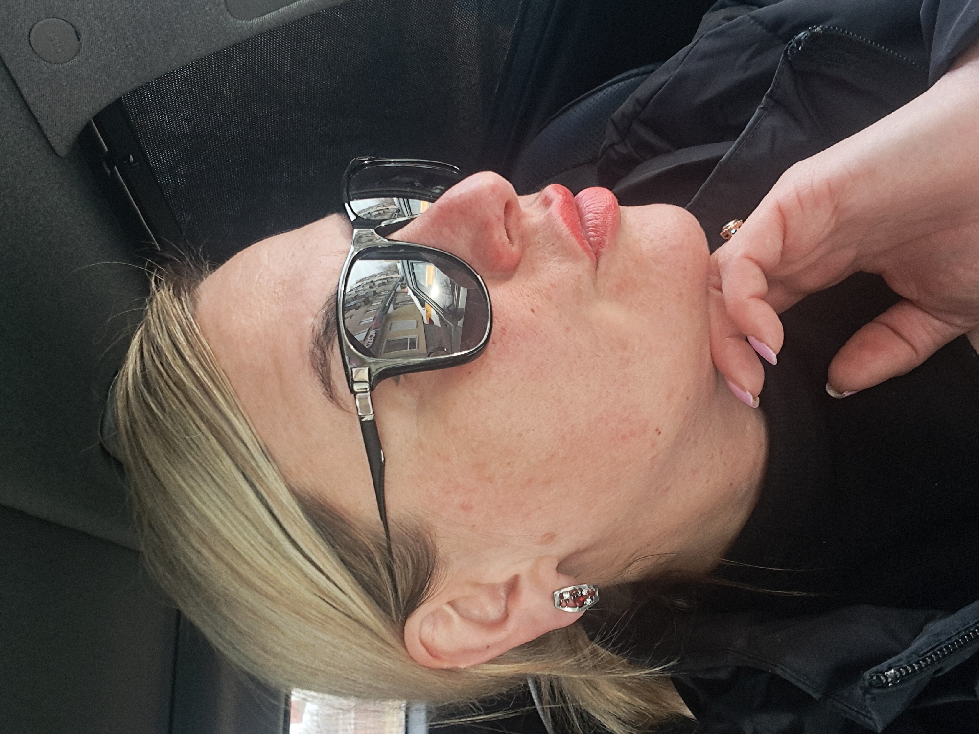 A blonde female, positioned in a vehicle, is depicted in profile view, facing towards the right side of the frame. Her hair is pulled back, with some loose strands visible. She wears large, black-framed sunglasses, which reflect a street scene featuring light-colored, multi-story buildings and parked cars, including a white sedan. Her left ear displays a silver-toned earring adorned with multiple small red and clear stones. Her lips are pursed, covered in pink-coral lipstick. Her right hand is raised to her chin, with fingers gently supporting her jawline; her fingernails are painted light pink. She is dressed in a black high-neck top under a black zippered jacket. The vehicle interior shows a dark gray fabric headliner and an exposed black mesh sunshade on the passenger side. A dark, possibly leather, seat is partially visible behind her. This scene takes place in Elektrostal, Russia.