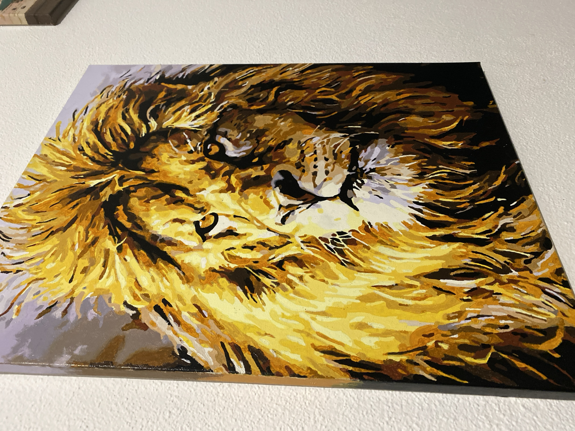 A close-up, low-angle shot captures a painting of a lion's head hanging on a textured white wall. The painting is rendered in a stylized, bold brushstroke technique, with dominant shades of yellow, orange, brown, and black. The lion's mane is depicted with vibrant, energetic strokes, framing its face. The lion's eyes are intense, with visible pupils and irises. The background of the painting is a soft gradient of purple and gray. The painting is mounted on the wall in Purkersdorf, Austria.