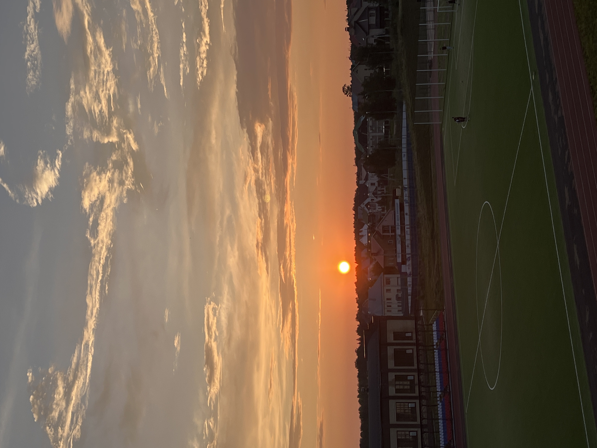 An aerial, high-angle shot captures a sunset over a sports field and residential area in Noviy, Russia. The sun, a bright orange orb, is low on the horizon, casting a warm glow across the sky, which is filled with wispy, illuminated clouds. Below the sky, a line of trees separates the sky from a collection of houses with red-tiled roofs and other residential buildings. A large, dark building with windows and bleachers is visible in the foreground, to the left of the frame. In front of this building is a green, artificial turf sports field with white markings, including a center circle and penalty box lines. A few figures, too small to discern details, are present on the field. The scene is framed by a dark red track on the bottom edge of the image.