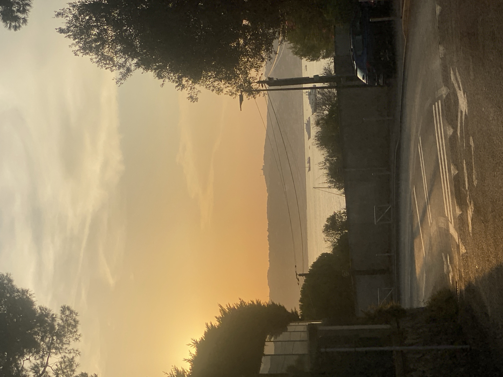 The image depicts a coastal view at sunset in Antibes, France. A road curves into the foreground, with white lane markings illuminated by the low sun. On the left, a building with glass windows is partially visible, nestled amongst lush green foliage. To the right, a dark-colored car is parked near a grey concrete wall. Beyond the wall, trees and bushes line the waterfront, and several boats are anchored in the calm sea. The distant hills are silhouetted against a hazy orange sky, with a few wispy clouds adding texture to the twilight. The overall impression is one of serene coastal living as day transitions into night.