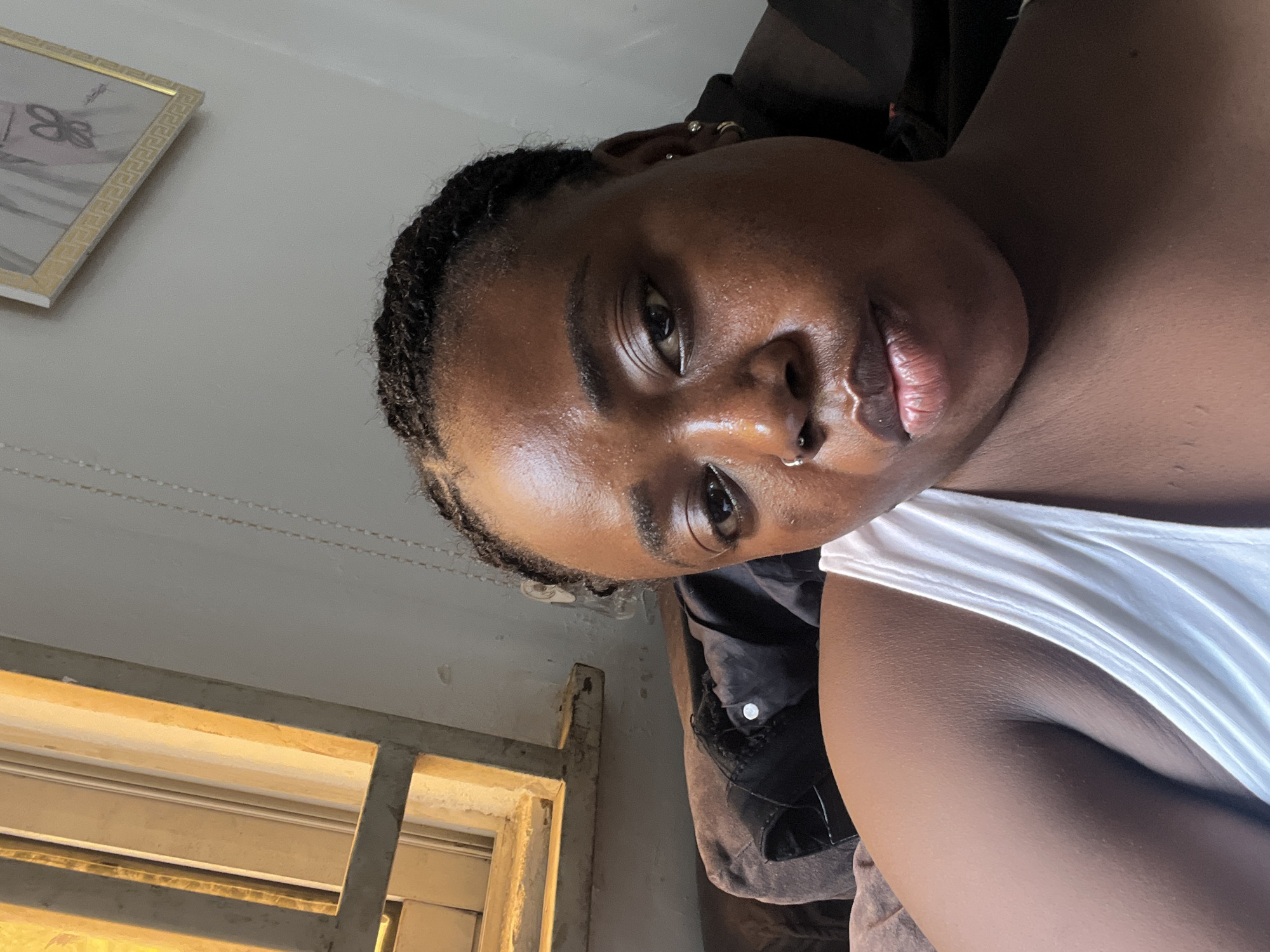This is a close-up, overhead shot of a young Black woman taking a selfie. She is looking directly at the camera, with her head tilted slightly. She has braided hair, a nose ring, and earrings. The background is a light-colored wall with a framed picture and a window with vertical blinds. The lighting suggests it is daytime. The overall mood is relaxed and casual. The woman is wearing a white tank top. The setting appears to be indoors, possibly a home, in Abuja, Nigeria. There is no visible text in the image.