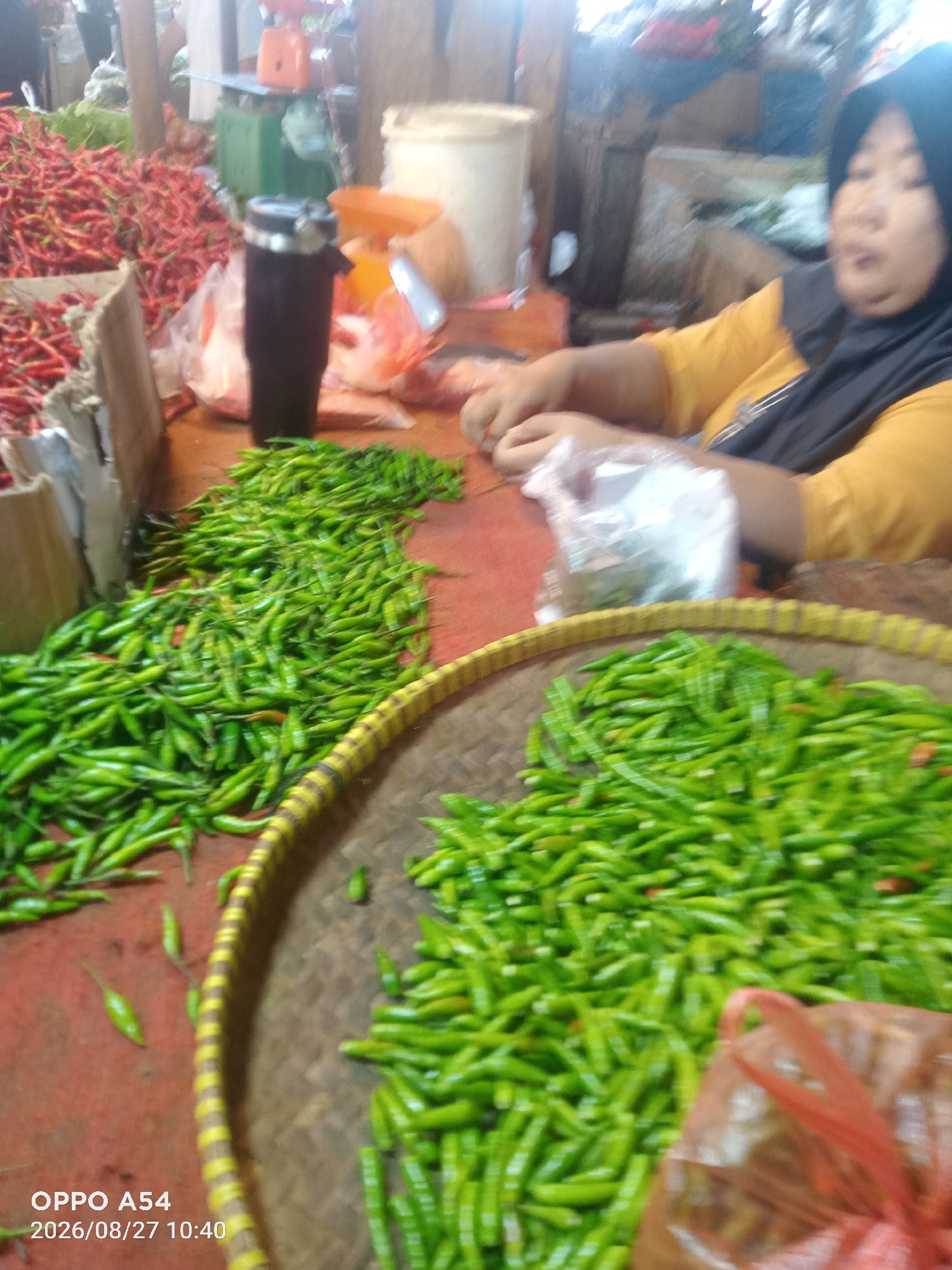 The image depicts a market scene, likely in Jambi, Indonesia, with a focus on a large display of green chili peppers. The chilies are spread across a red surface and also piled into a woven basket with yellow and brown stripes. To the left, a significant amount of red chili peppers is visible, forming a mound. A black cylindrical object, possibly a thermos or container, stands on the red surface near the red chilies. In the background, a woman wearing a yellow shirt and a black hijab is partially visible, engaged in an action that appears to be sorting or handling something, with her hands near a clear plastic bag. Various market stalls and structures are blurred in the background, suggesting a busy marketplace environment. The overall lighting is somewhat diffused. The bottom left corner of the image displays text indicating "OPPO A54" and a date and time stamp.