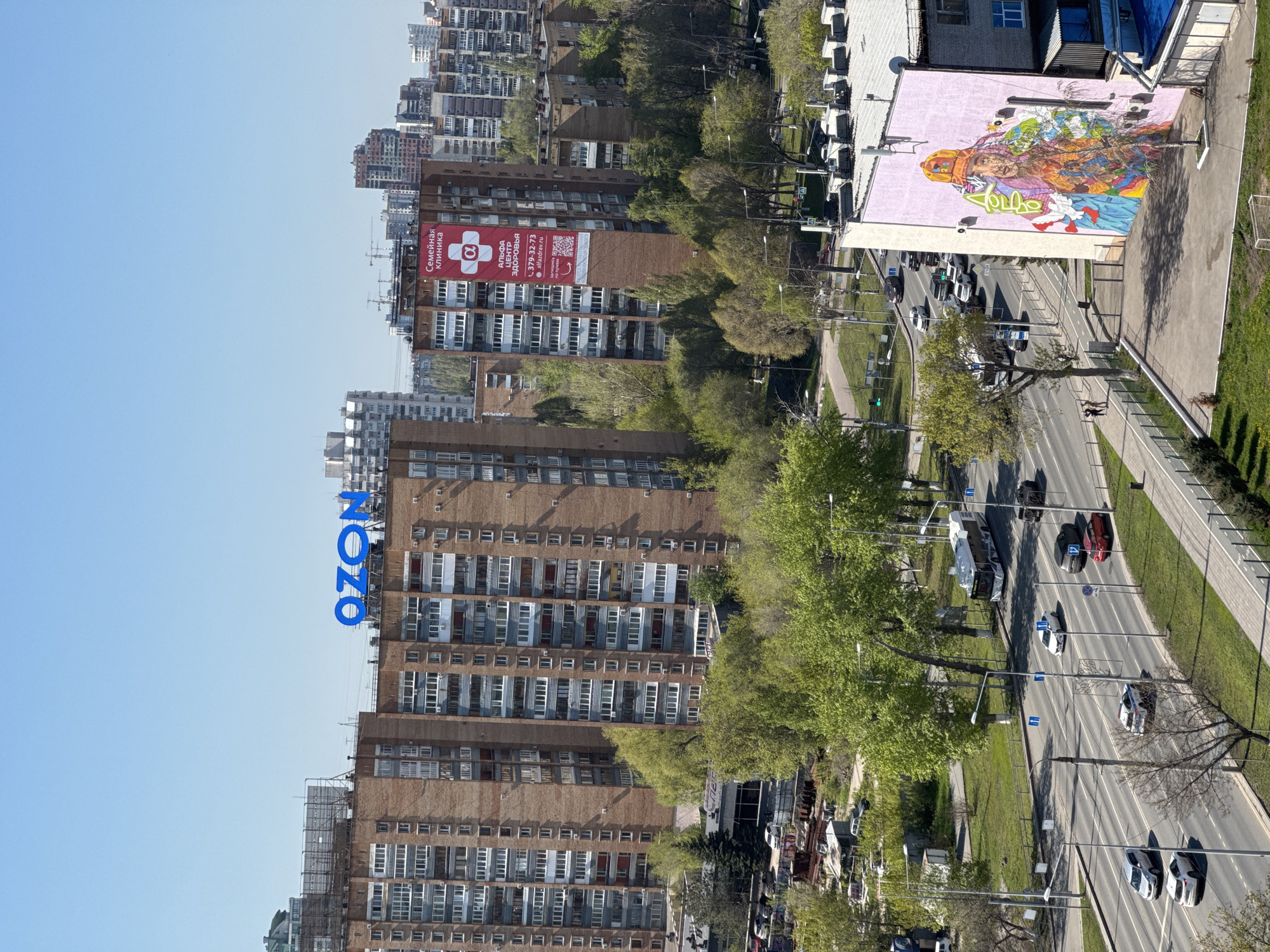 A medium shot of an urban area on a sunny day. Tall brick apartment buildings dominate the left side of the image, with the blue "OZON" logo prominently displayed on one. Banners with advertisements are seen on the buildings. The right side of the image features a street with cars driving on it, a sidewalk, grass, and trees lining the street. A building with a large mural is visible on the right side of the street. Pedestrians are seen walking on the sidewalk.