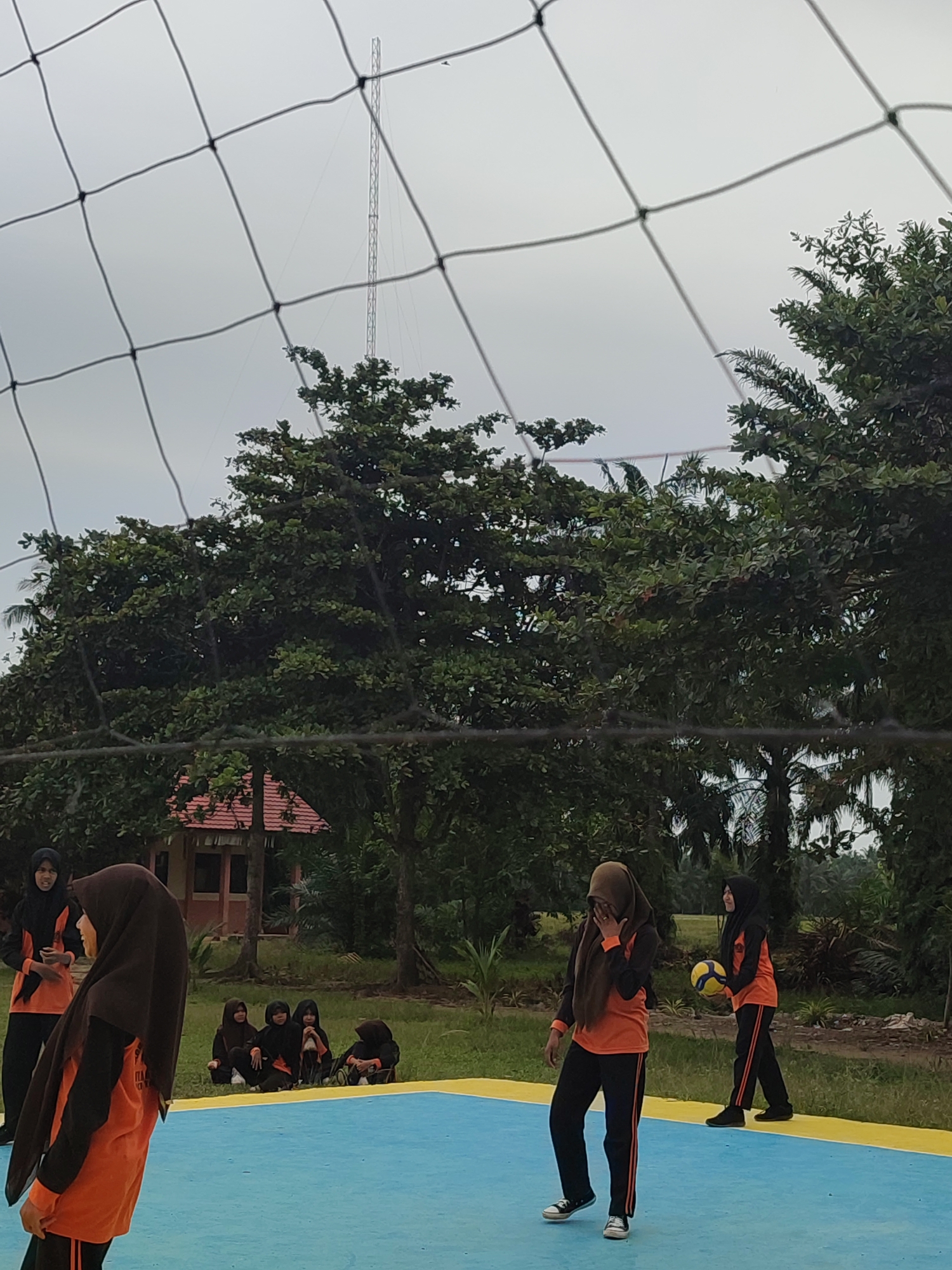 In Blangriek, Indonesia, on a cloudy day, a group of young women are playing volleyball on an outdoor court with a blue and yellow surface. The court is bordered by grass and surrounded by trees. A volleyball net stretches across the foreground, obscuring parts of the scene. In the background, a red-roofed building and a tall radio tower are visible. Several women are on the court, one in the foreground on the left, wearing an orange shirt and brown hijab, facing away from the camera. Another woman in the foreground on the right, also in an orange shirt and brown hijab, is holding a volleyball and looking towards the left. In the middle ground, several women are sitting on the grass, wearing hijabs and casual clothing. The overall atmosphere appears to be one of casual sports activity in an outdoor setting.