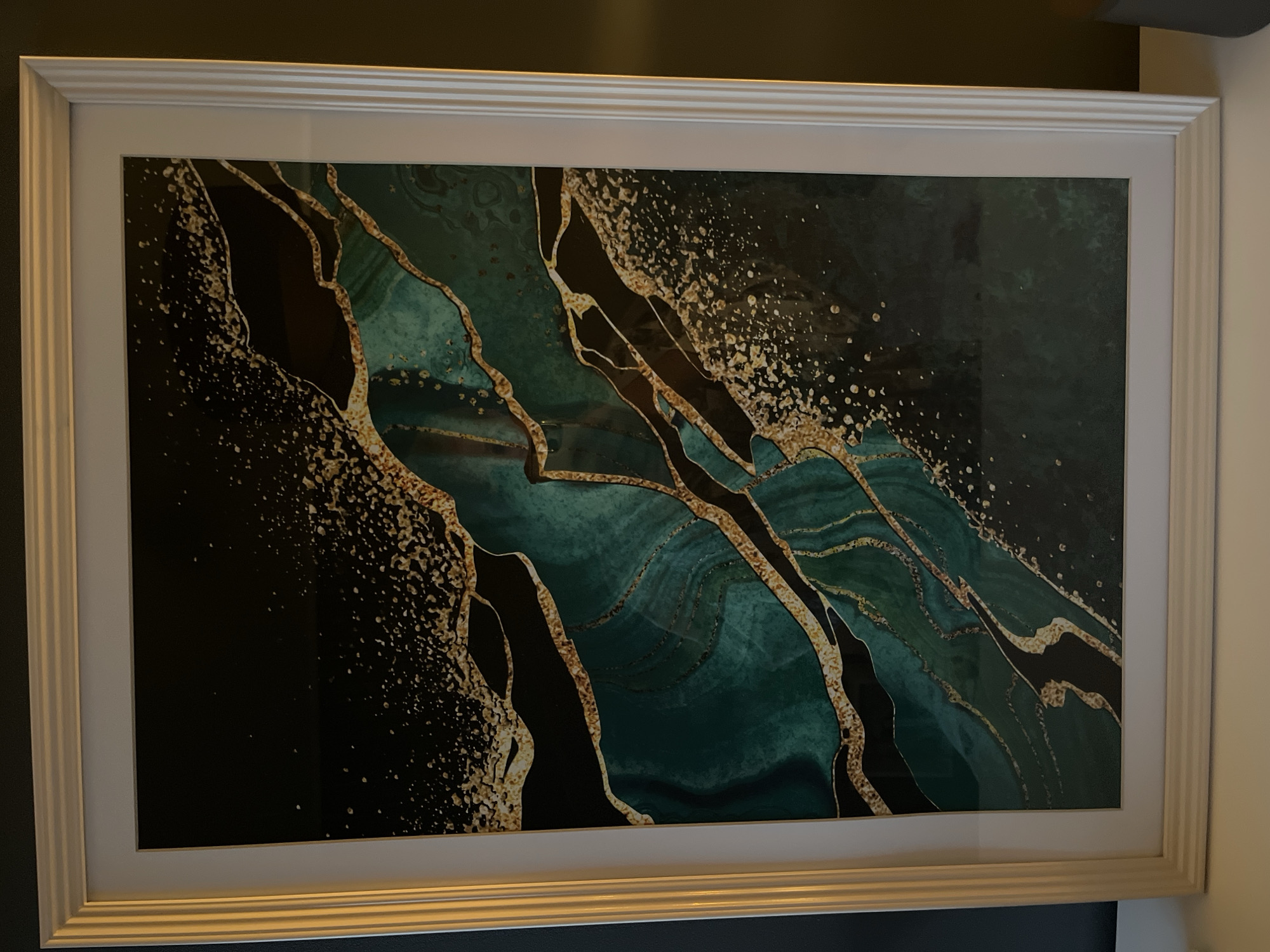 A framed abstract artwork with a white frame is displayed on a shelf. The artwork itself features a dark background with swirling patterns of deep teal and black, interspersed with golden, glitter-like accents and thin, gold-lined organic shapes. The scene appears to be indoors, possibly in a domestic setting in Haabneeme alevik, Estonia. The lighting is dim, casting a subtle glow on the artwork's frame. No people are visible in the image.