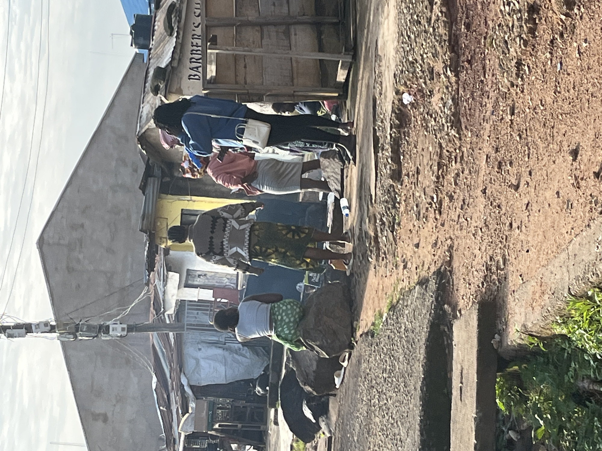 The image depicts a street scene in Jos, Nigeria, featuring several individuals and the urban environment. In the foreground, a person is seated on large rocks, facing away from the camera, wearing a white tank top and a green patterned skirt. To their right, another individual stands, wearing a patterned top and a long, green patterned skirt. Further to the right, two more individuals are visible. One, wearing a pink top and a gray skirt, is facing partially towards the camera, while the person to their left, in a blue jacket, faces away, holding a phone to their ear and carrying a white purse. Behind them, a wooden structure with "BARBER'S C..." visible suggests a barbershop. The background reveals a building with a corrugated metal roof, typical of informal settlements, and a utility pole with multiple meters attached. The ground is uneven, with patches of gravel and dirt. The overall scene suggests a typical day in a residential or commercial area in Jos.