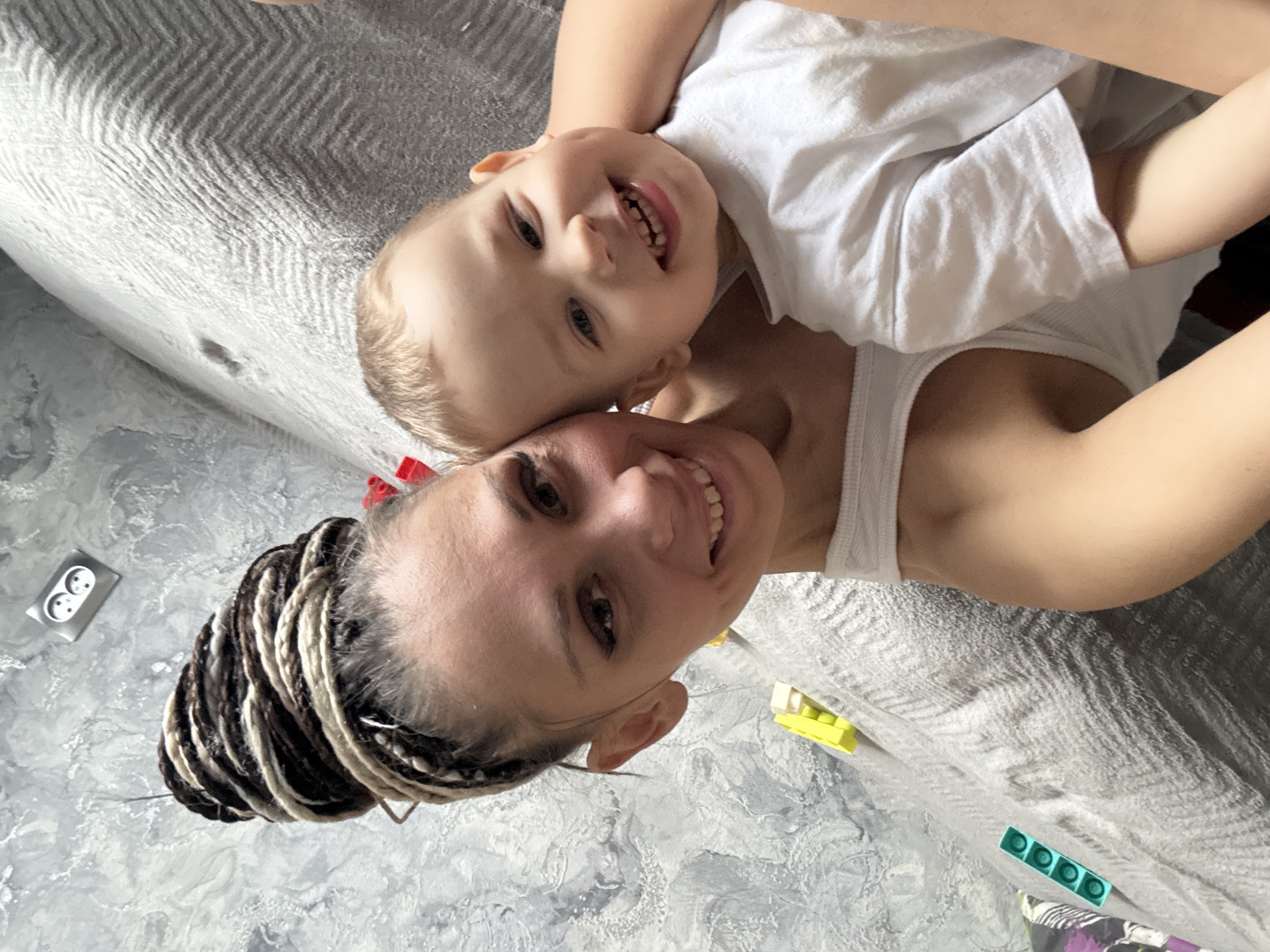 The image depicts a woman and a young child in a close embrace, likely indoors. The woman has dark hair styled in braids with light blonde highlights, fair skin, and is wearing a white tank top. She is smiling and looking directly at the camera. The child, also fair-skinned, is wearing a white t-shirt and is positioned partially on top of the woman. The child is also smiling, and its teeth are visible. They are lying on a gray textured blanket or bedspread. In the background, portions of a wall are visible with an electrical outlet and what appear to be children's toys, specifically Lego bricks, scattered nearby. The image appears to be taken indoors, likely at home, judging by the intimate nature of the scene and the domestic environment. There is no specific indication that it is taken in Yaroslavl', Russia or any other city.