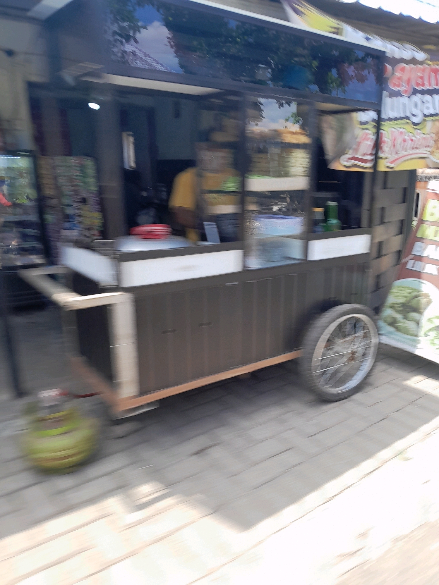 The image shows a blurred street scene in Jatirejoyoso, Indonesia, with a food cart as the central subject. The food cart is dark brown with a white countertop and a metal wheel on the right side. A green gas cylinder is visible on the left, also blurred. The background is also blurry, but appears to be a shop or building with signage. A person wearing a yellow shirt is partially visible behind the cart. The ground is paved with light-colored rectangular stones, and the overall lighting suggests it is daytime. The blur indicates motion, likely from the photographer or the cart itself.