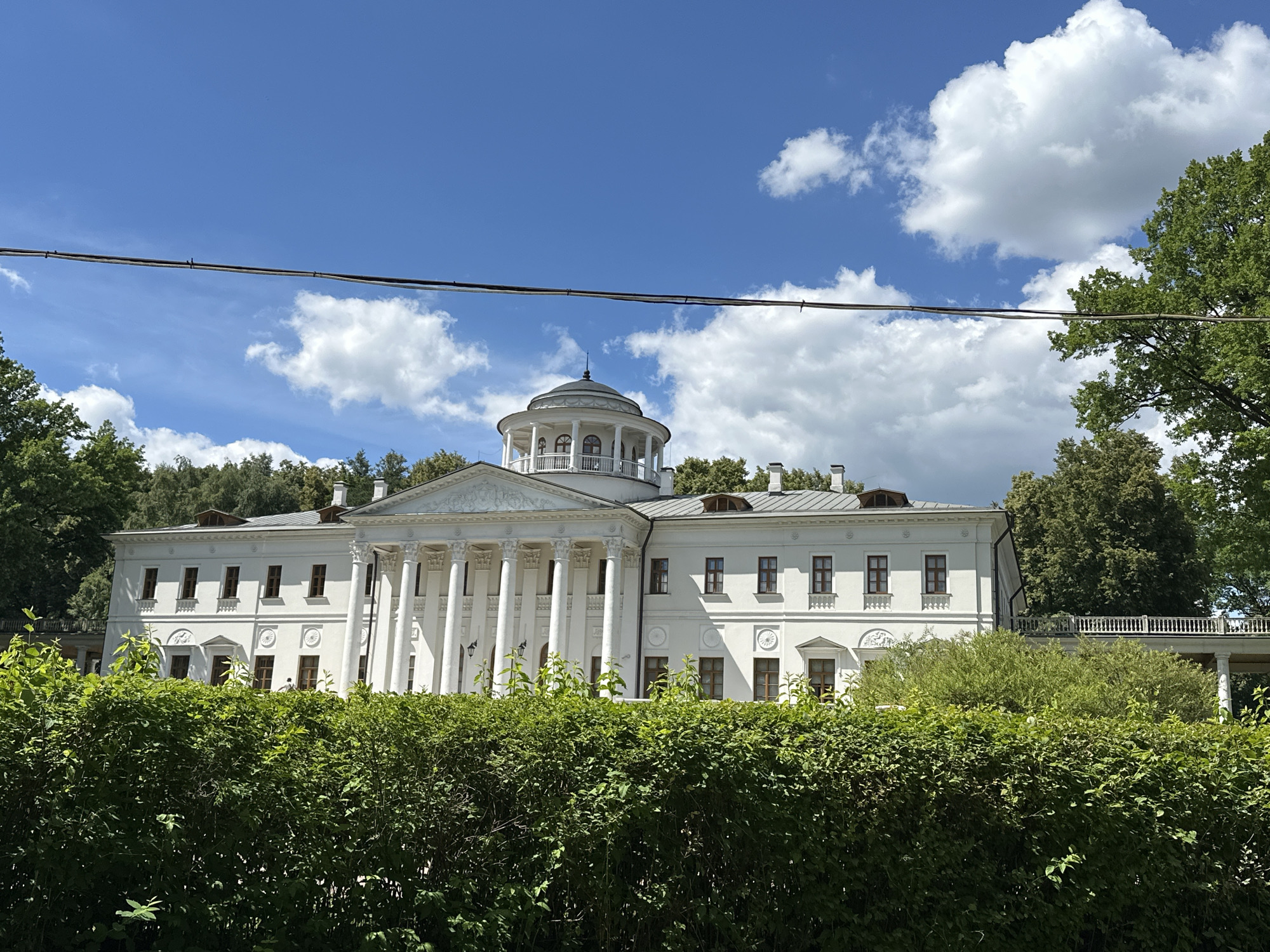 The image shows a white neoclassical building with a circular dome on top, surrounded by lush green trees and a dense hedge in the foreground. The building features a prominent portico with Corinthian columns and a pediment. Several windows are visible on the facade, some with decorative elements above them. A balcony with a white railing extends from the right side of the building. The sky is blue with scattered white clouds. Power lines are visible across the top of the image. The scene is the estate of Ostafievo, located in the village of Shcherbinka in the Novomoskovsky Administrative Okrug of Troitsky and Novomoskovsky Administrative Okrugs of Moscow, Russia, which was formerly part of the Moscow Oblast.