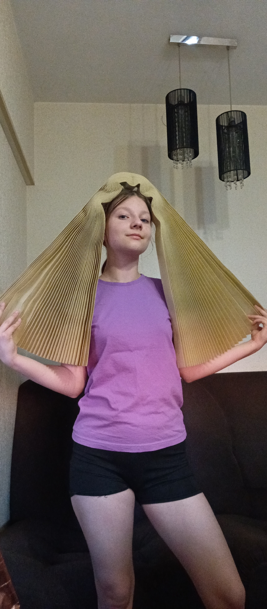 A young woman stands indoors, wearing a lavender T-shirt and black shorts. She has a decorative, pleated, golden-hued object on her head, which she holds up with both hands. Behind her is a dark brown couch. Overhead, two pendant lamps with black shades hang from the ceiling. The location is in a residential setting of an unknown city, with neutral-toned walls.